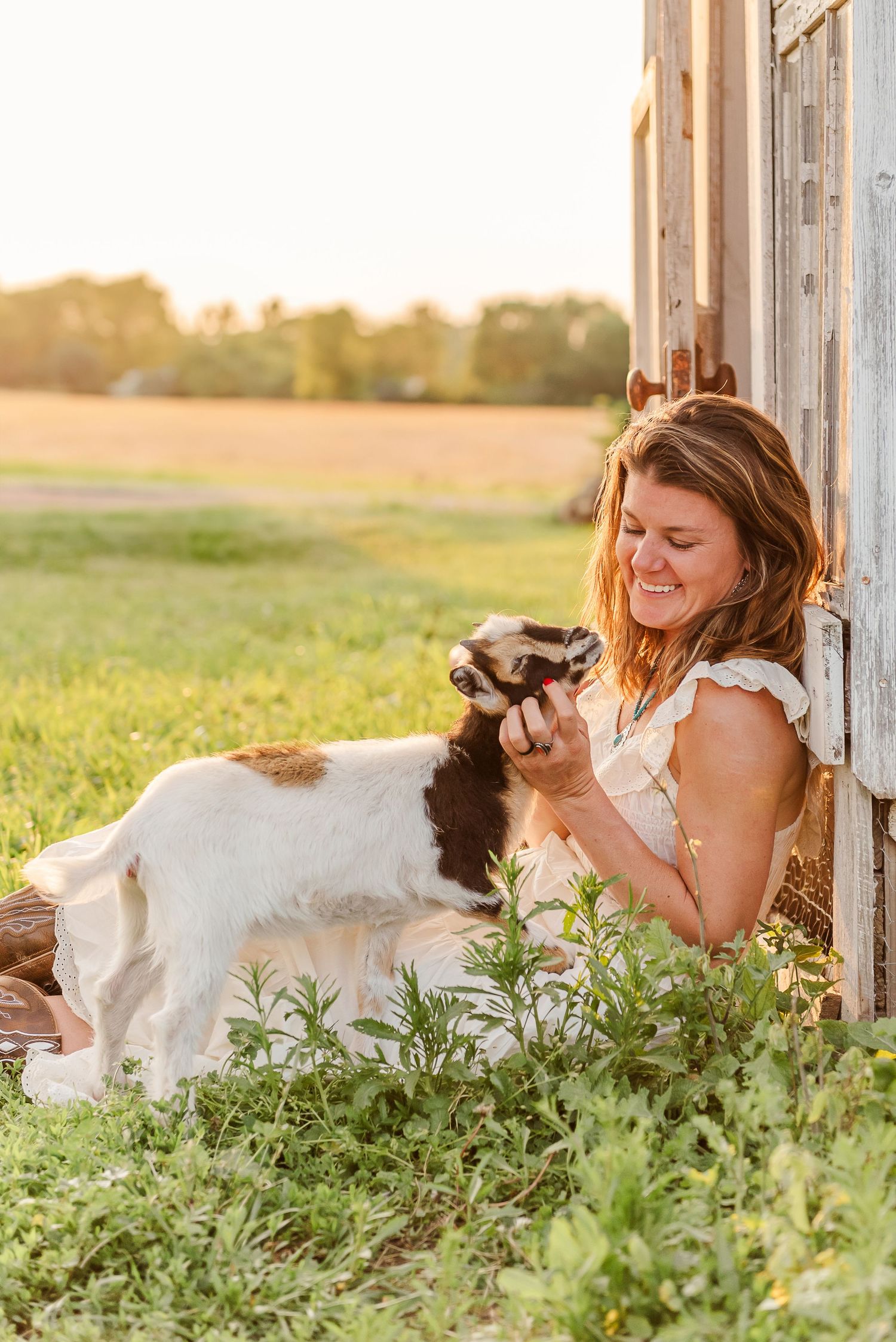 Woman in white dress laying in grass playing with baby goats during golden hour.