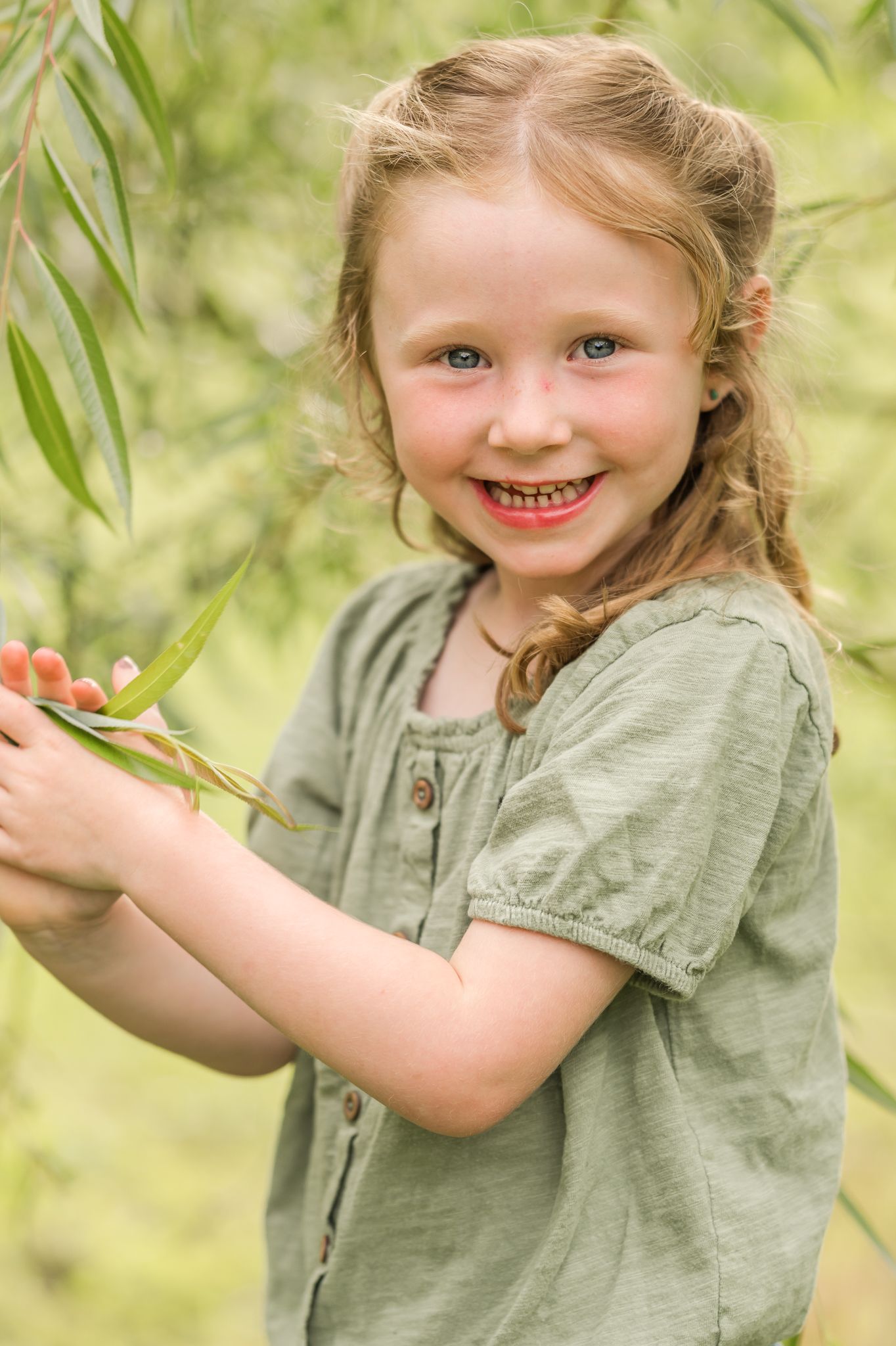 A smiling young girl in a green dress holds willow branches outdoors, her rosy cheeks glowing in natural light.
