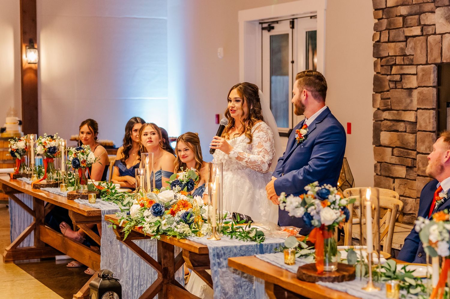 Wedding guests enjoy dinner at a rustic stone-walled reception venue with warm lighting and floral centerpieces.