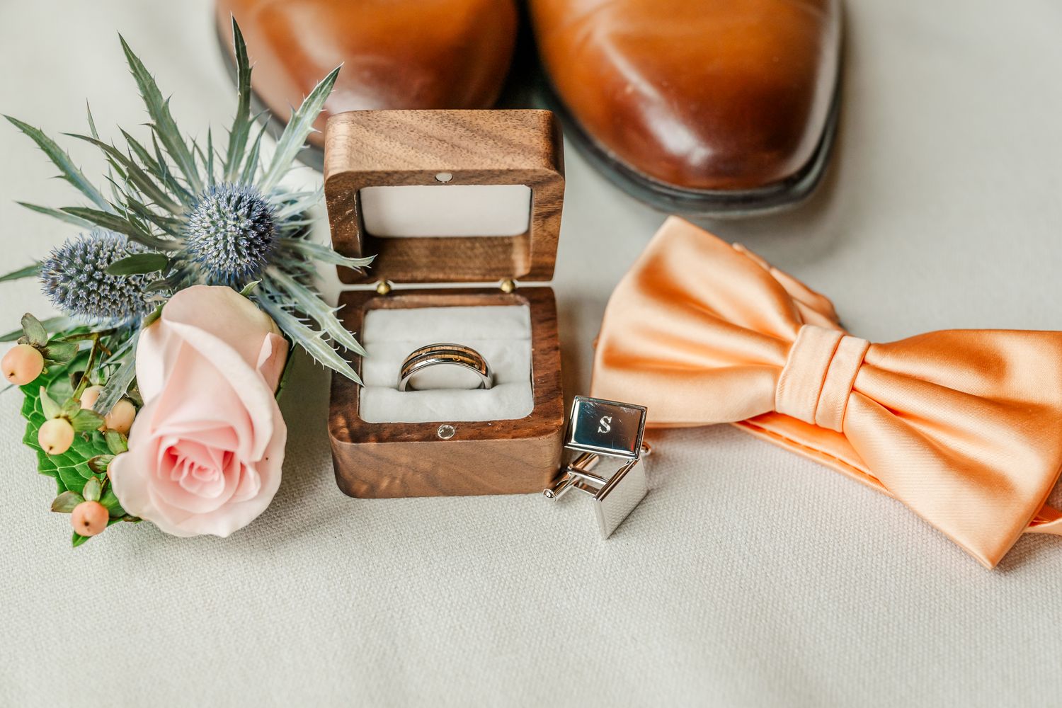 Wedding flat lay with gold ring in wooden box, peach bow tie, cufflinks, brown dress shoes, and pink rose bouquet.