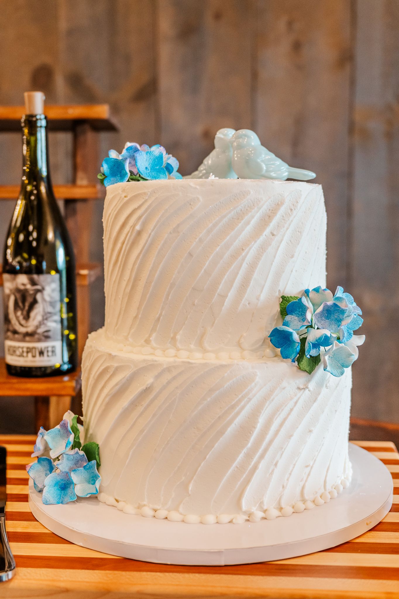 Two-tier white wedding cake with blue hydrangeas and dove topper, displayed near wine bottles on wooden table.The Old Barns at Dry Run Farms