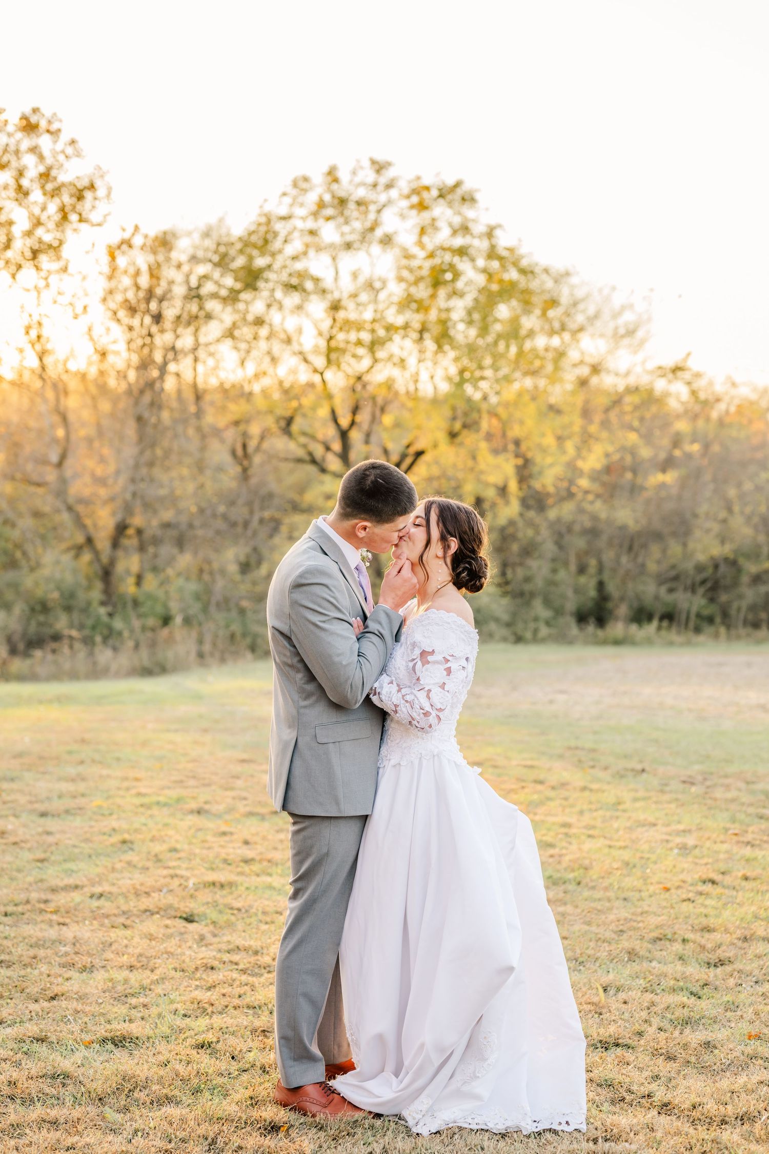 Romantic wedding couple embraces in a golden field at sunset with autumn trees in the background. Fall wedding photography.
