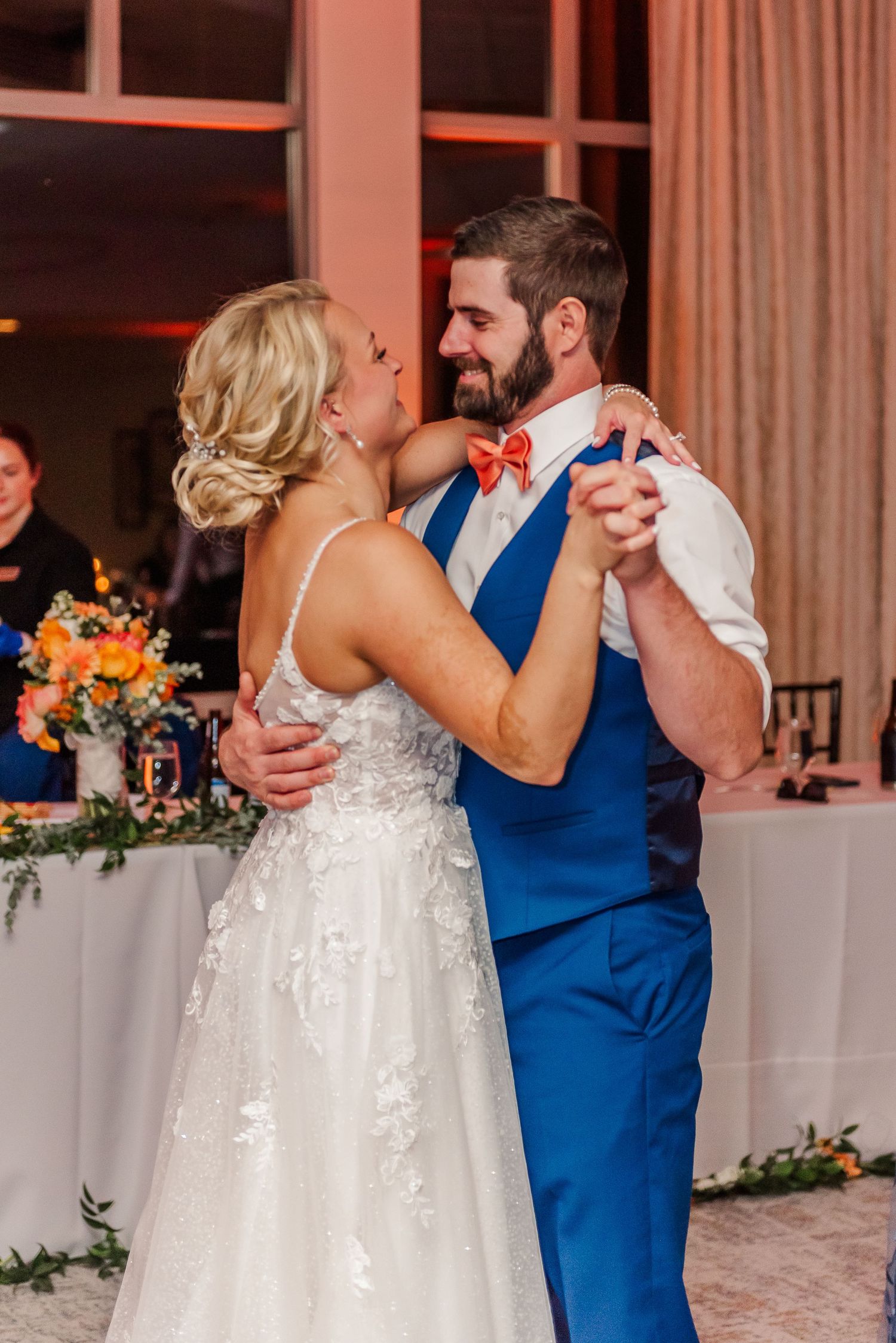 A couple dances together at a wedding reception, with the bride in white and partner in a blue vest.
