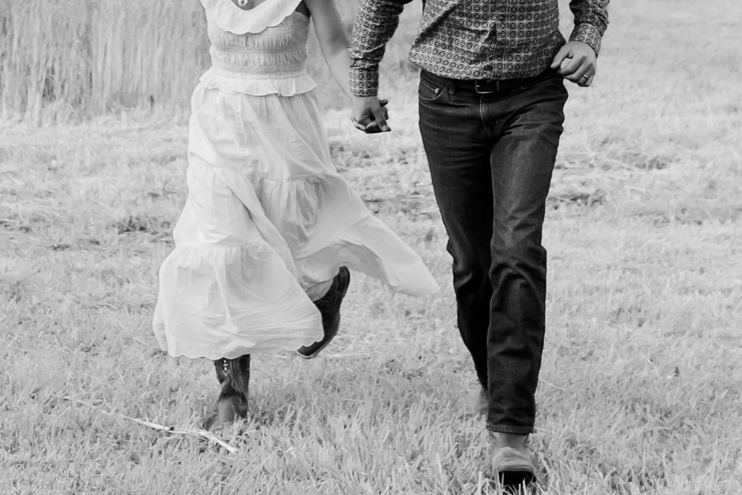 Black and white photo of two people walking through a field, one in a flowing white dress and cowboy boots.
