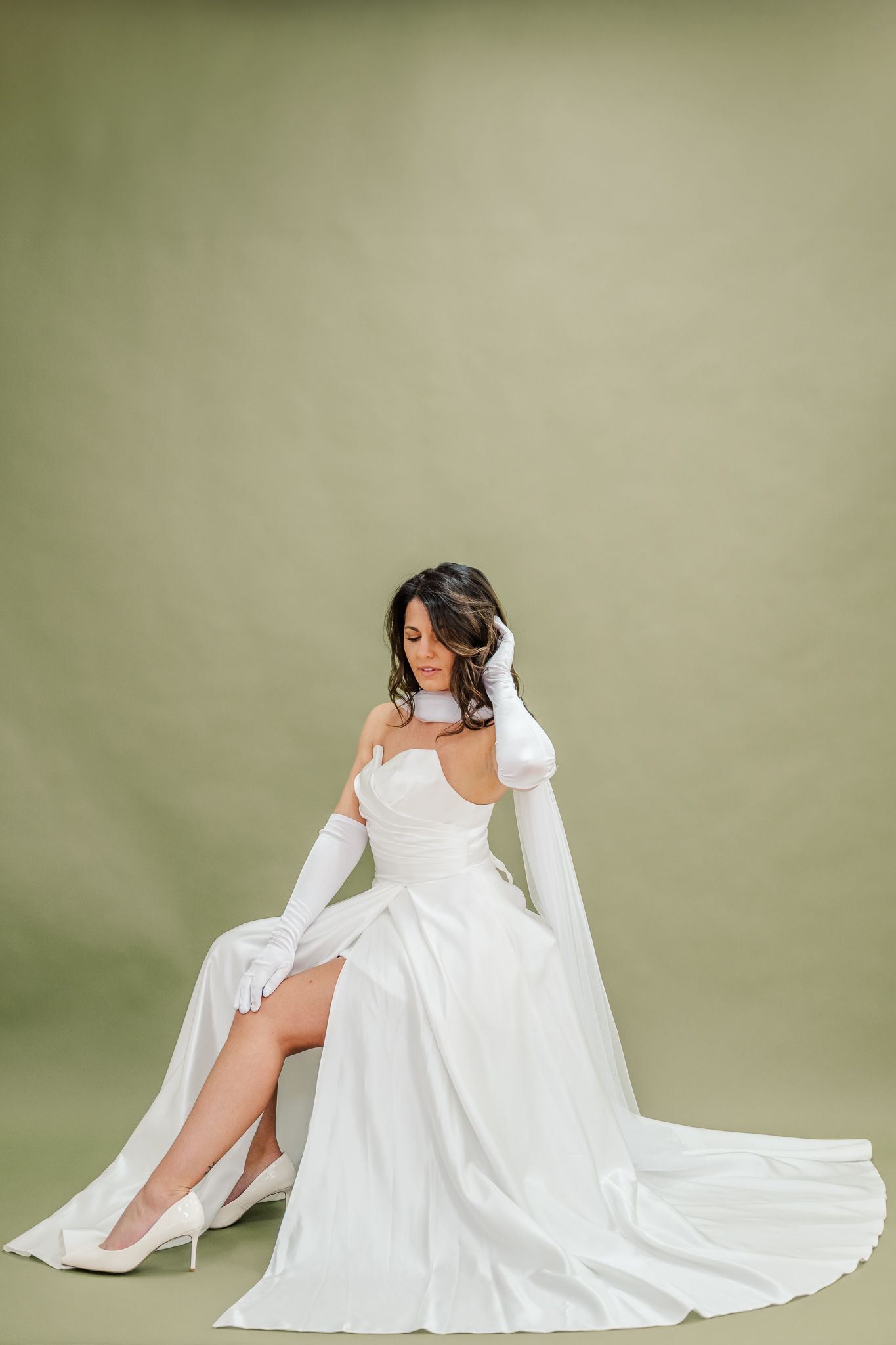 Elegant bride in white strapless gown with long gloves sits gracefully against sage green background.