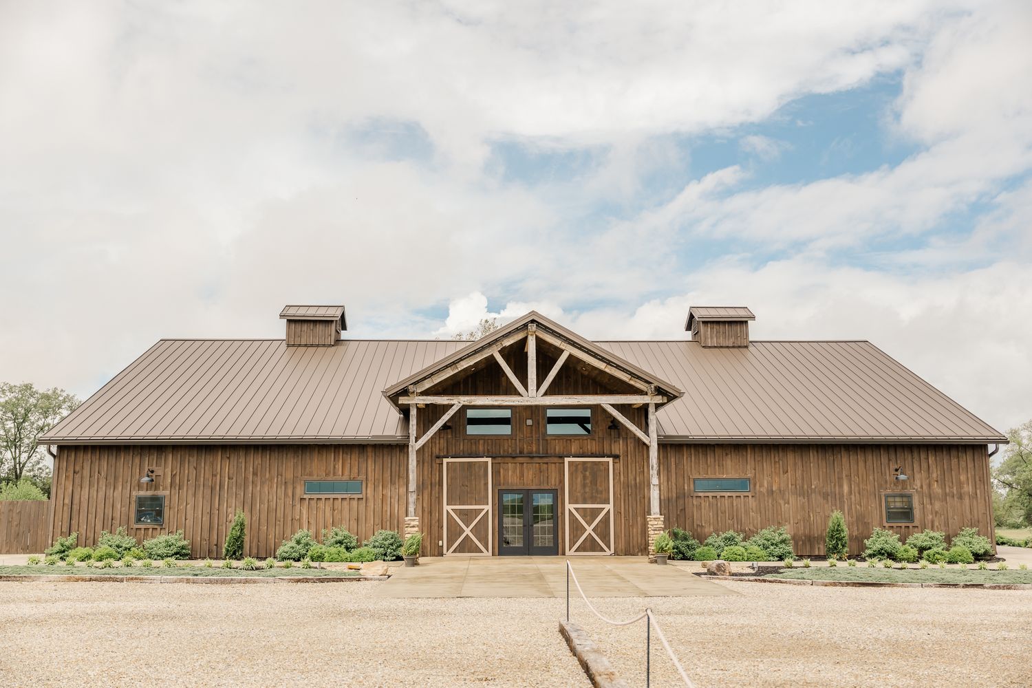 The Old Barns at Dry Run Farms venue with metal roof, timber frame entrance, and manicured landscaping under cloudy skies.