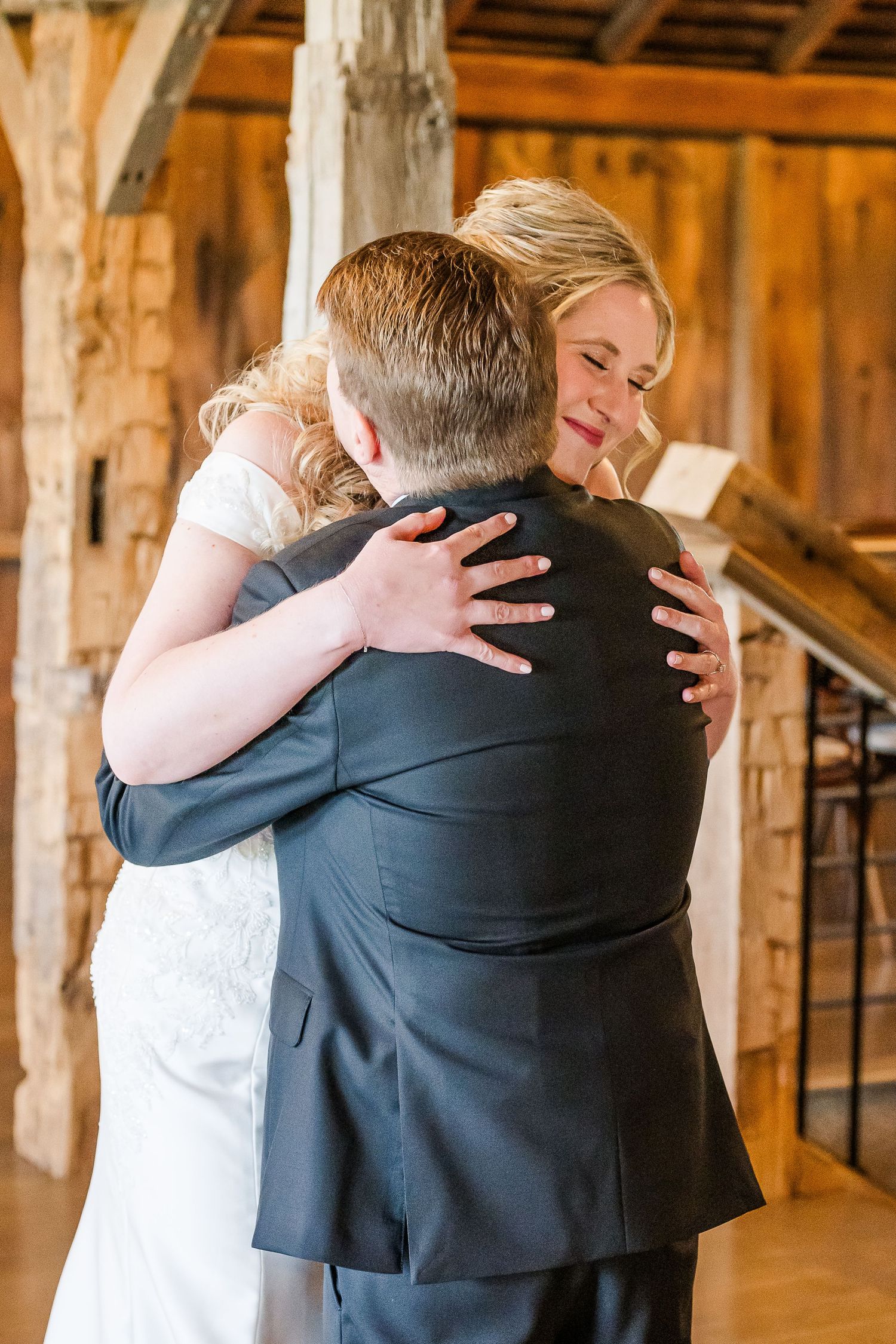 A tender embrace between a bride in white and groom in dark suit inside a rustic wooden barn venue.The Old Barns at Dry Run Farms