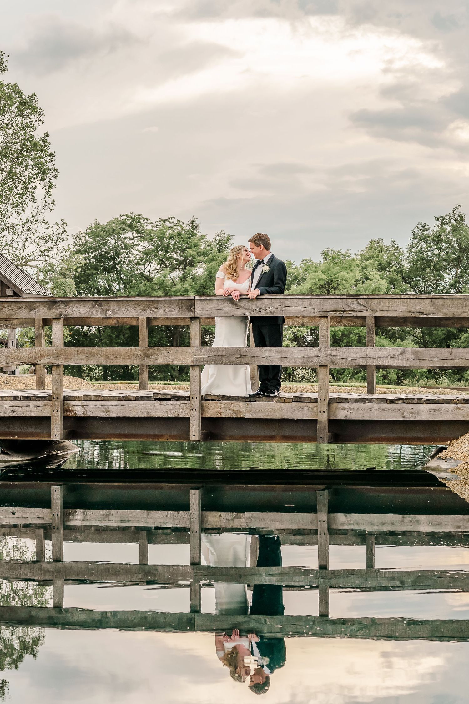 Wedding couple reflected in still pond water with wooden bridge railing.The Old Barns at Dry Run Farms in Williamsport, OH.