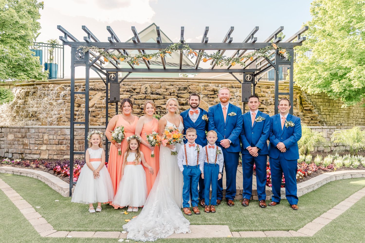 Wedding party poses under pergola with bridesmaids in coral and groomsmen in blue suits.