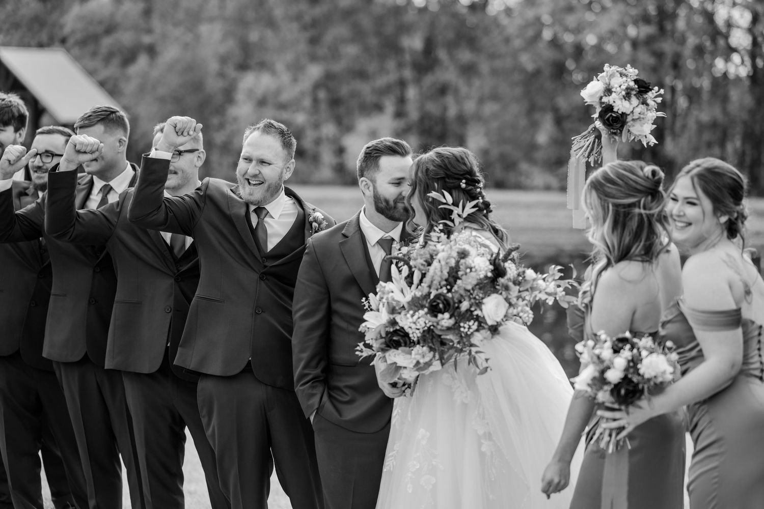 Wedding party members gather outdoors holding purple and white floral bouquets during a celebration.