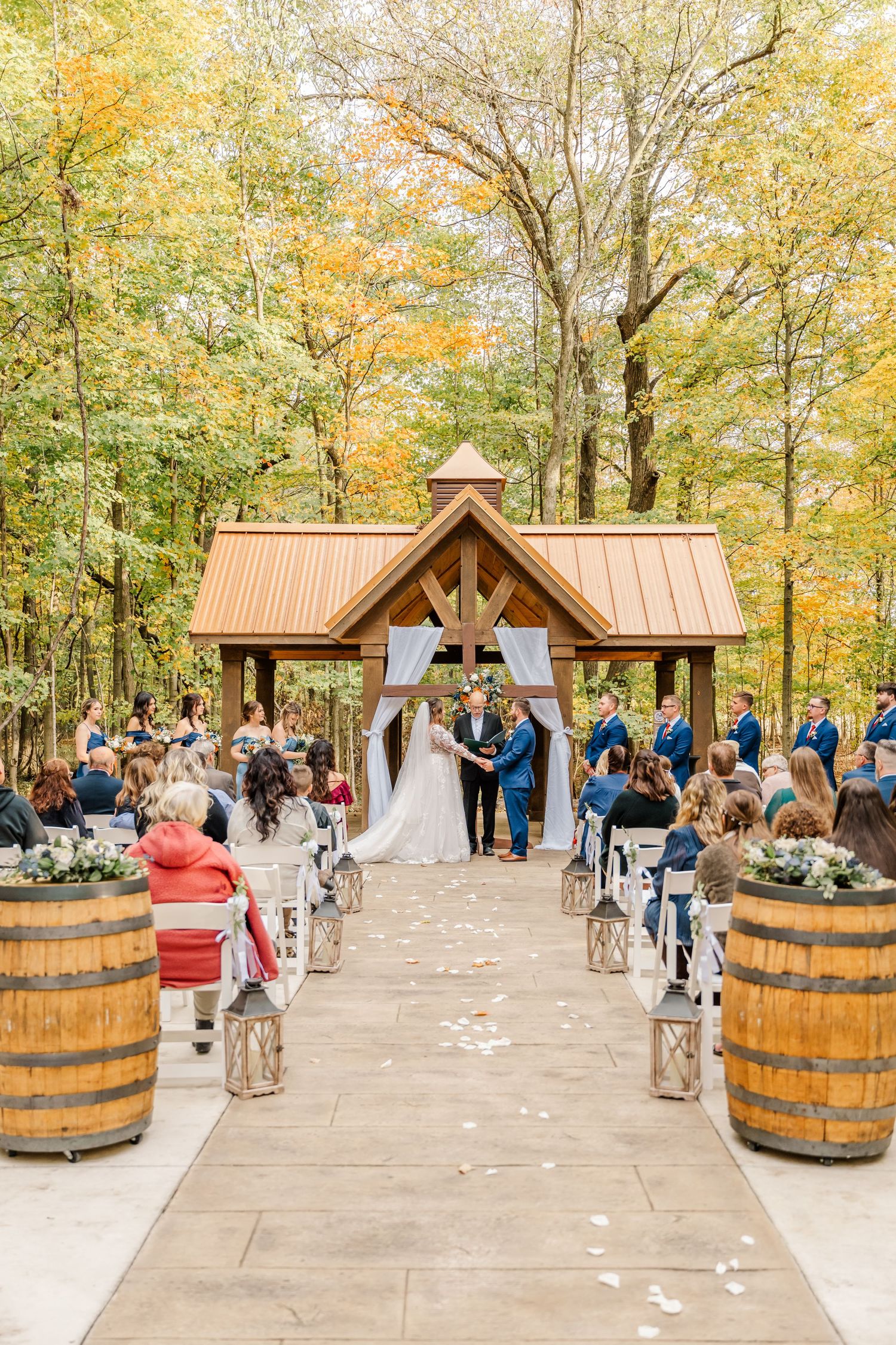 Rustic outdoor wedding ceremony under wooden pavilion with wine barrel decor and autumn foliage in background.Four Seasons Barn Wedding