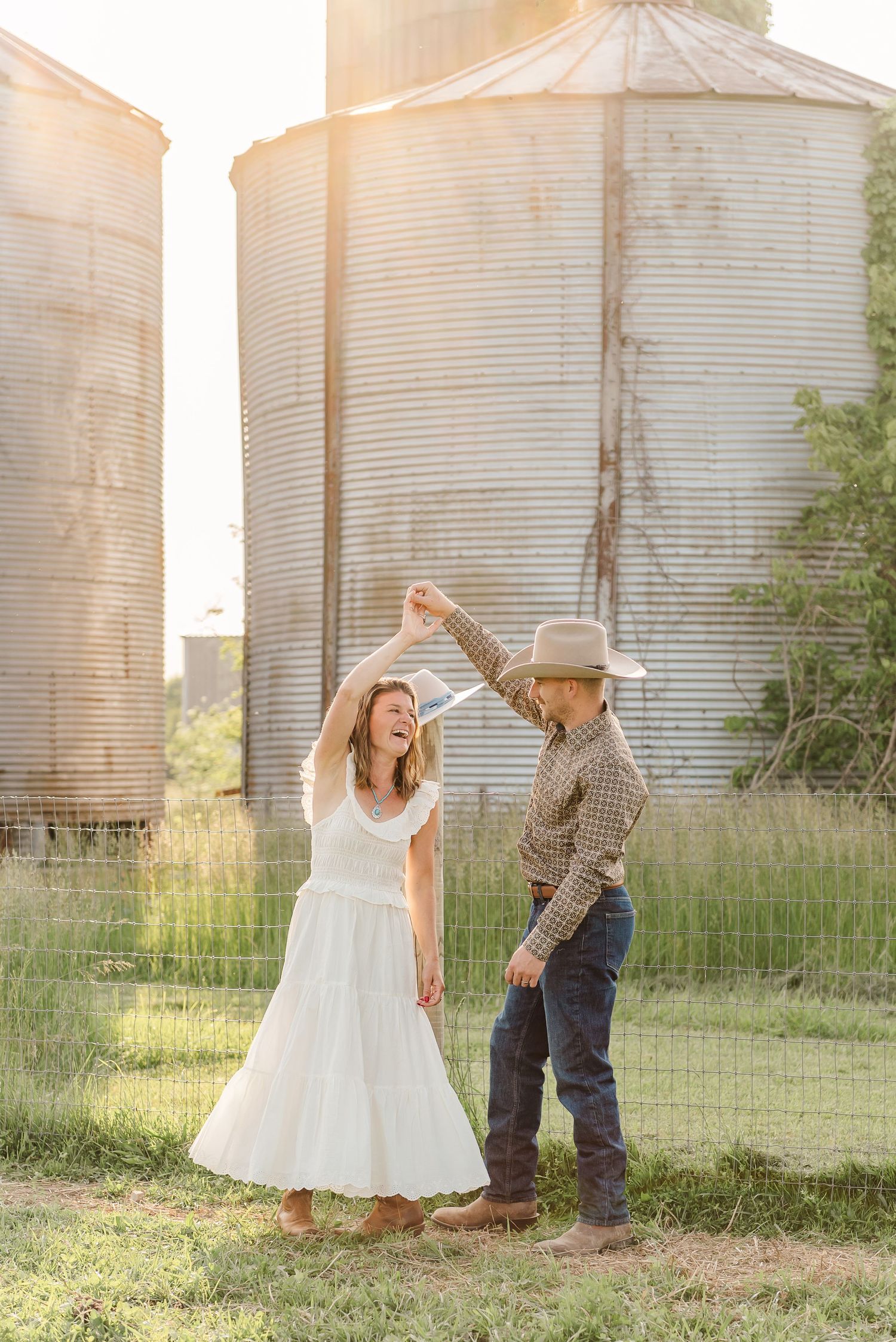 A romantic country photoshoot with a couple dancing and posing in front of metal grain silos at sunset.