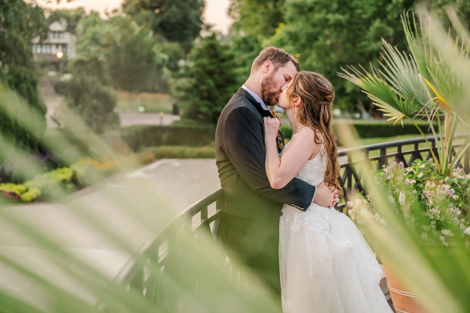 A romantic couple embraces in a garden setting with palm fronds in the foreground during golden hour.