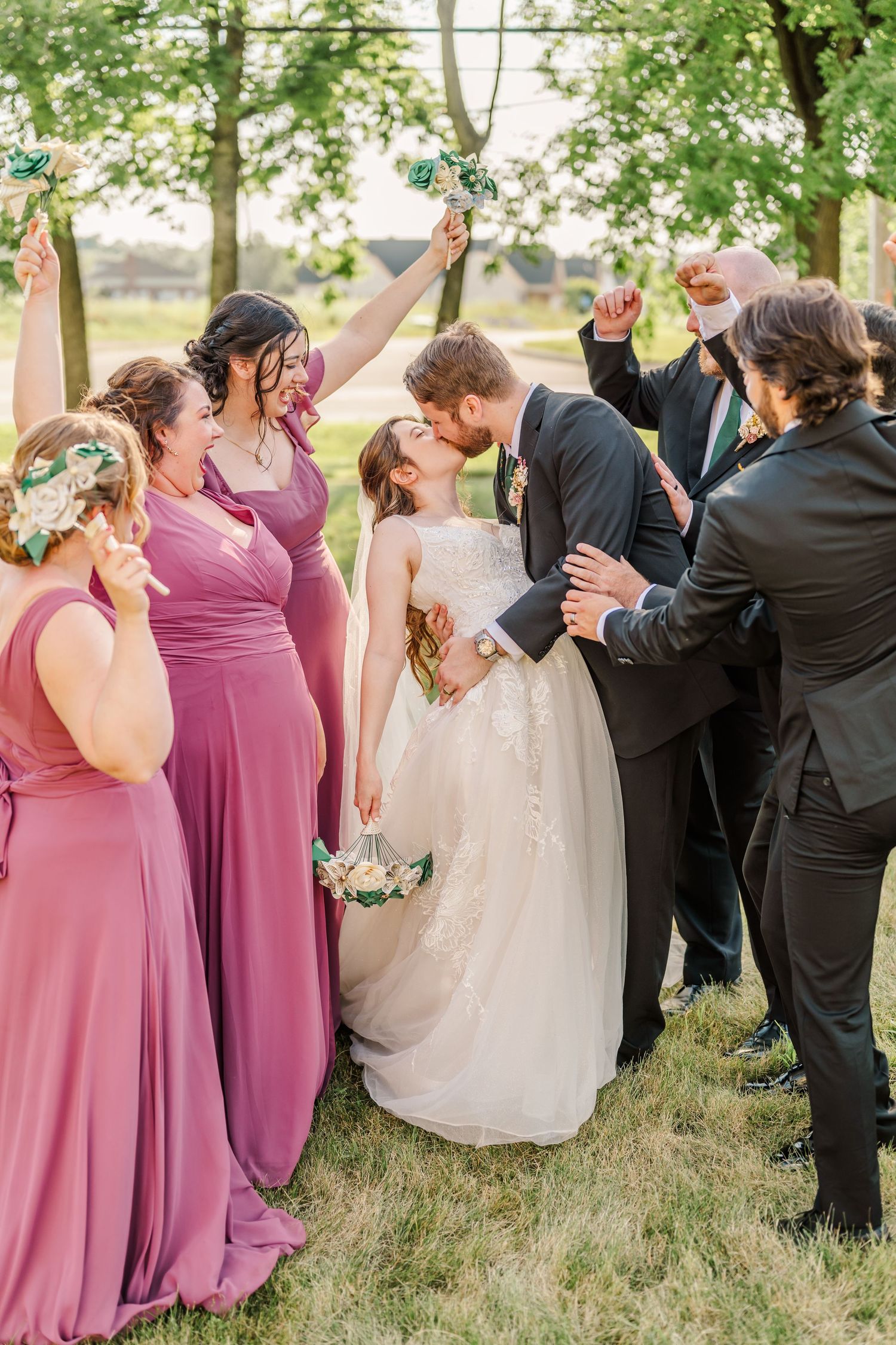 Wedding party celebrates outdoors as newlyweds share a romantic moment surrounded by bridesmaids in dusty rose dresses.