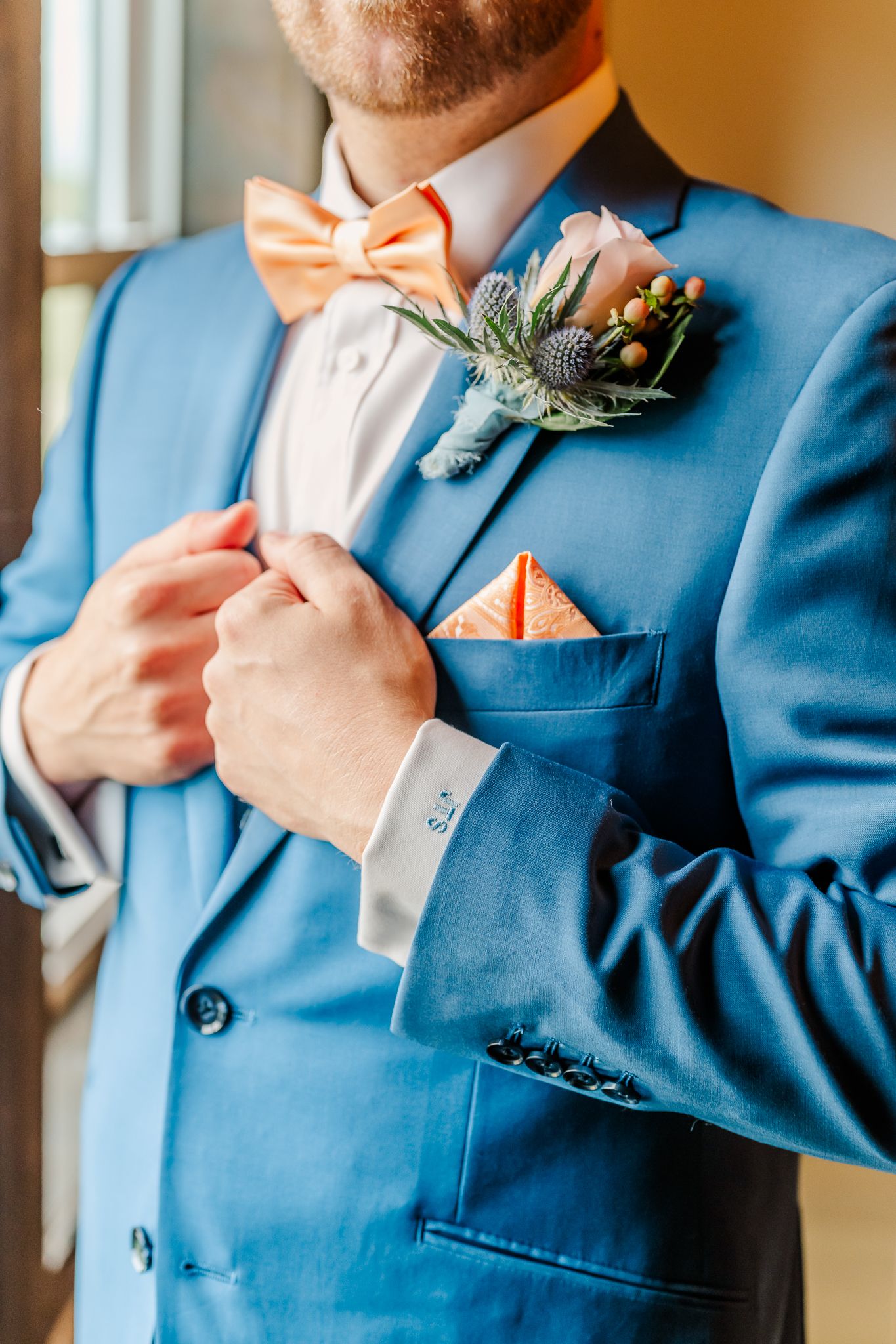 A groom in a blue suit adjusts his floral boutonniere, wearing an orange bow tie and matching pocket square.The Old Barns at Dry Run Farms.