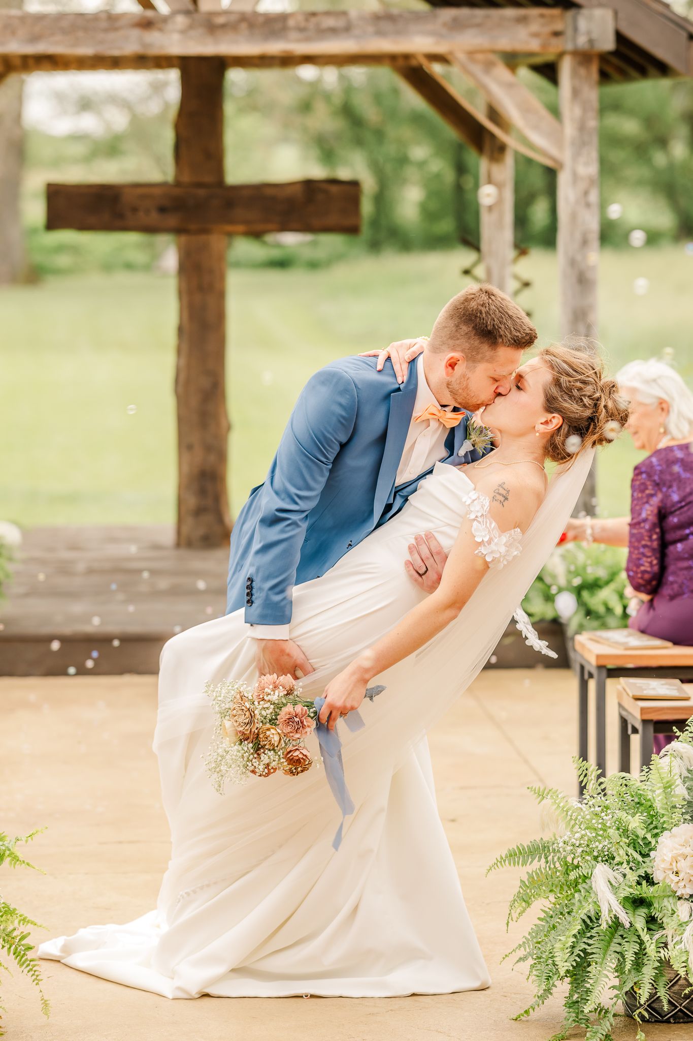 Groom dips bride for first kiss at rustic outdoor wedding ceremony near wooden cross. The Old Barns at Dry Run Farms in Williamsport, Ohio.