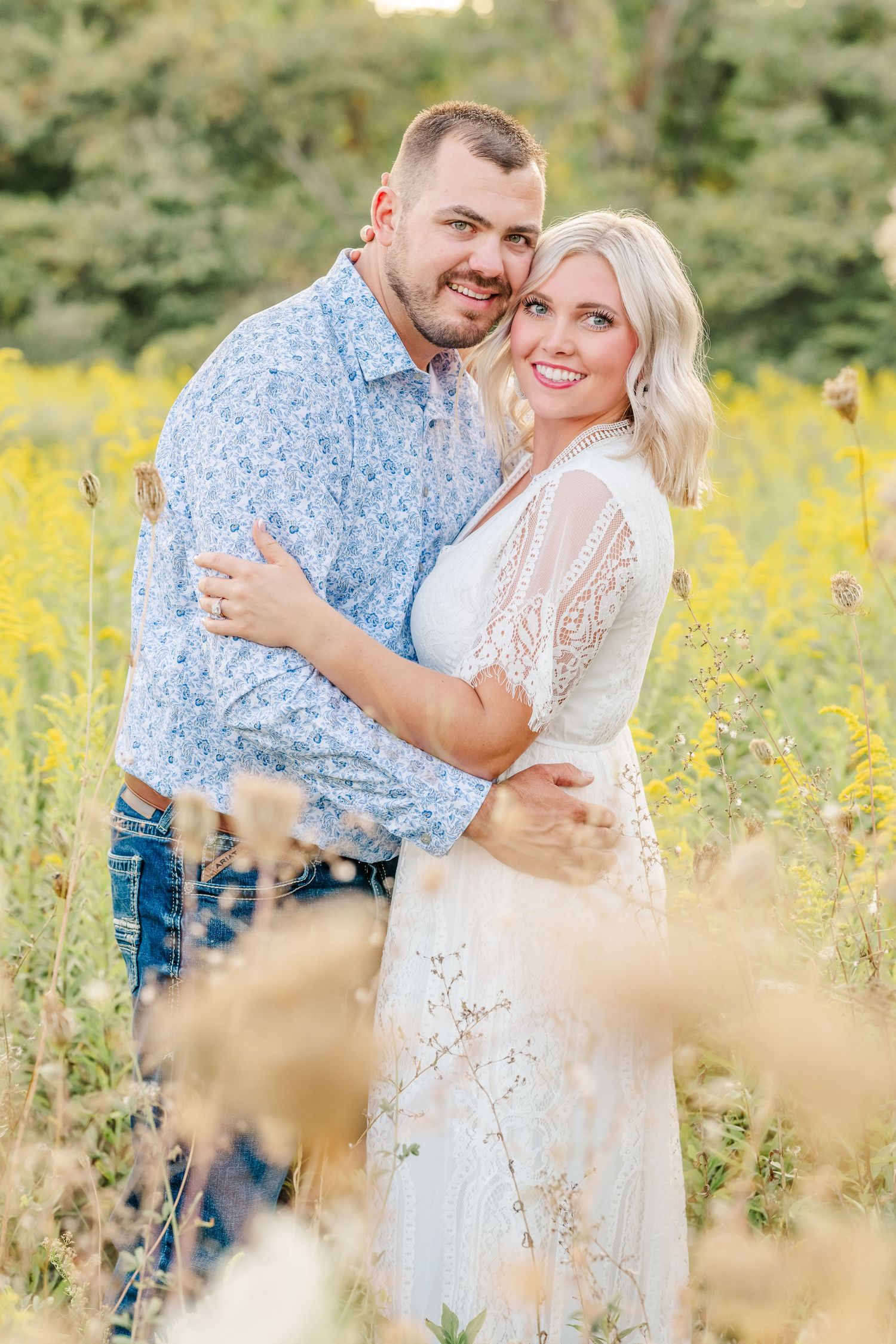 A couple embraces in a field of yellow wildflowers on a sunny summer day wearing white and blue outfits.