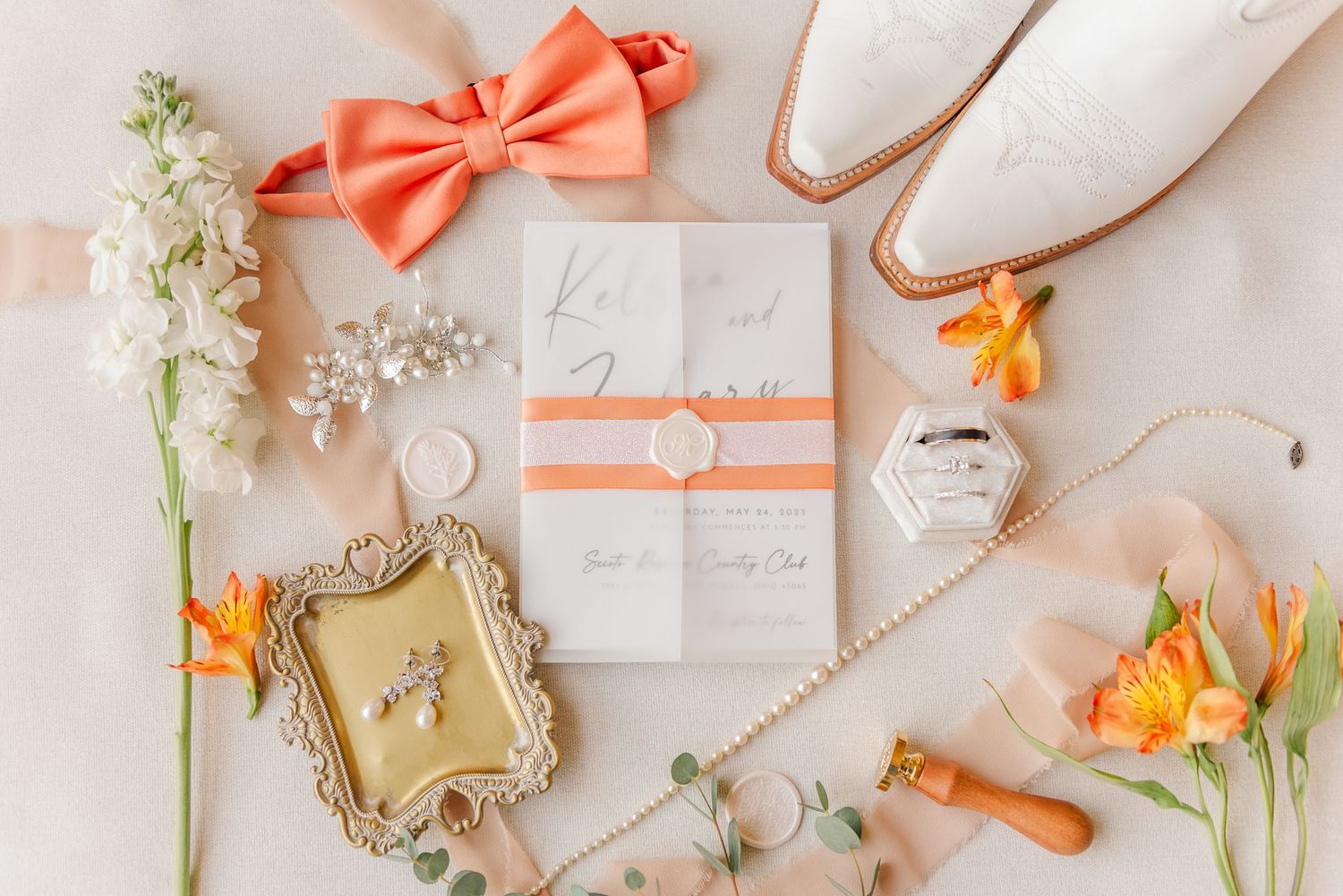 Wedding details flatlay with coral bow tie, white flowers, orange lilies, rings and invitation on beige background.