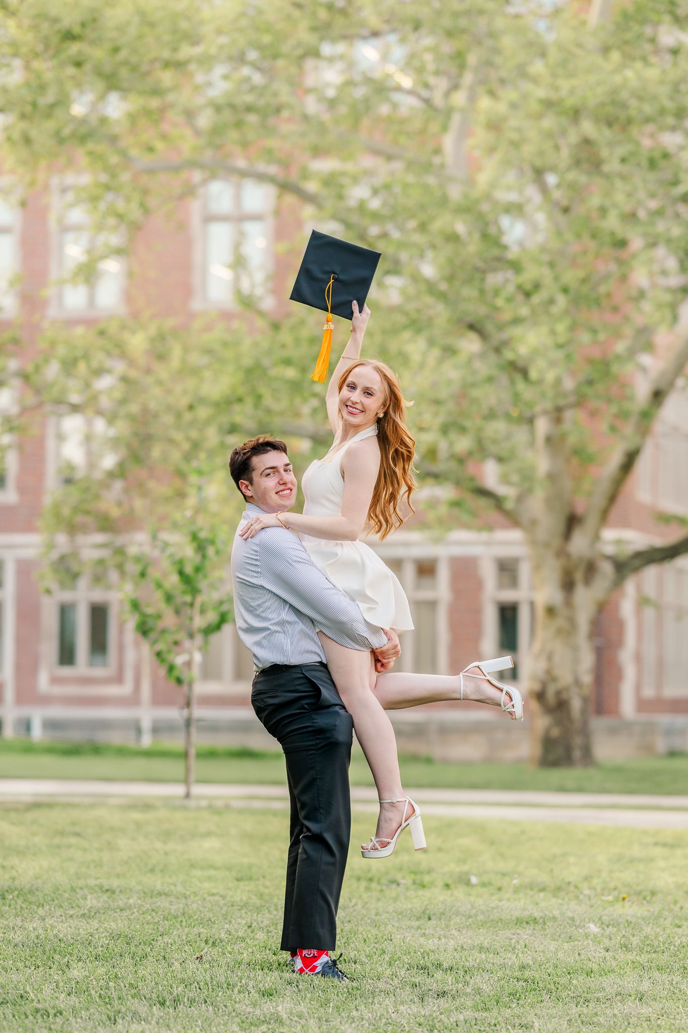 A young man lifts a smiling woman in a white dress holding a graduation cap outdoors on a green campus lawn. OSU campus.