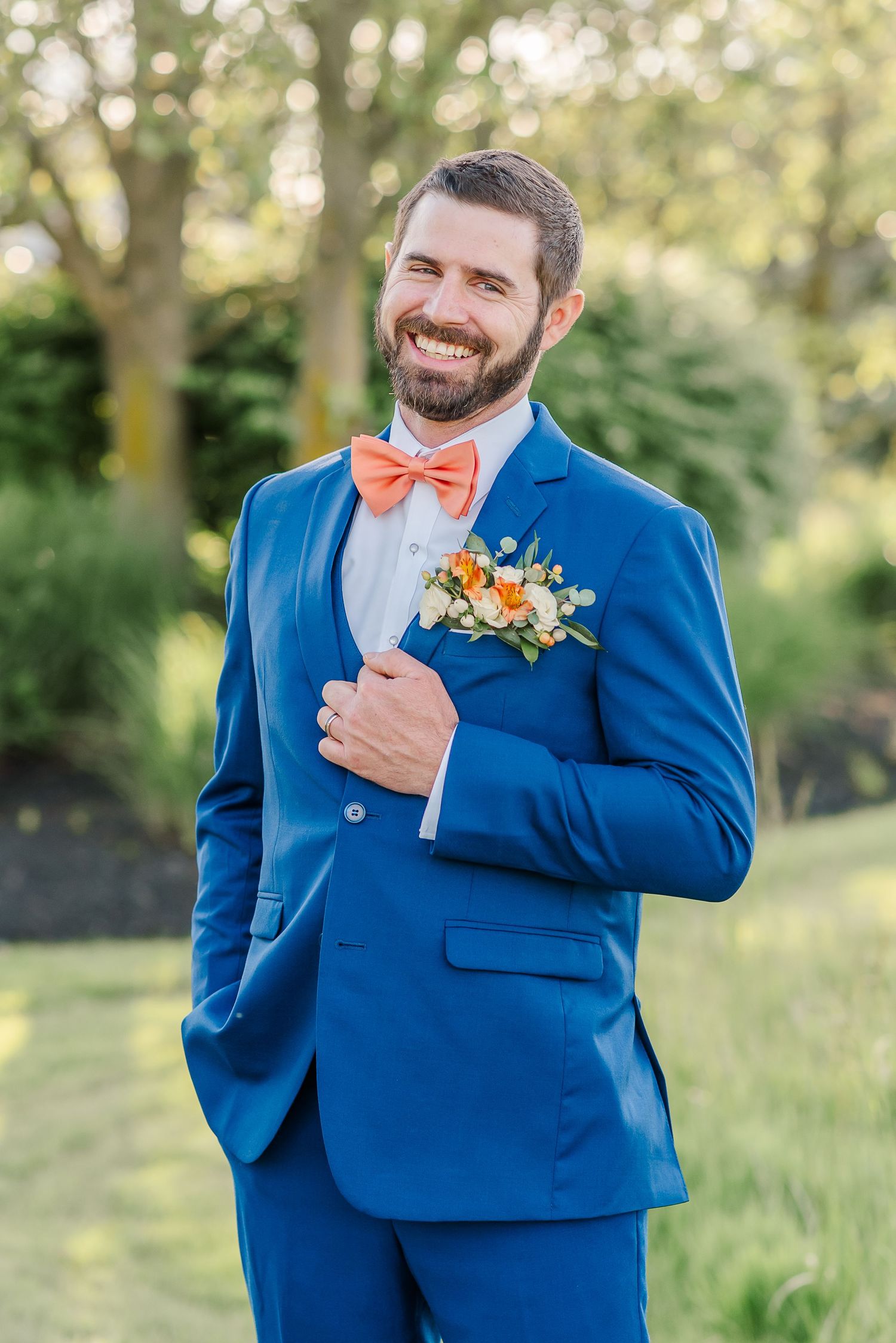 A groom in a bright blue suit with coral bow tie poses in a sunny outdoor setting.