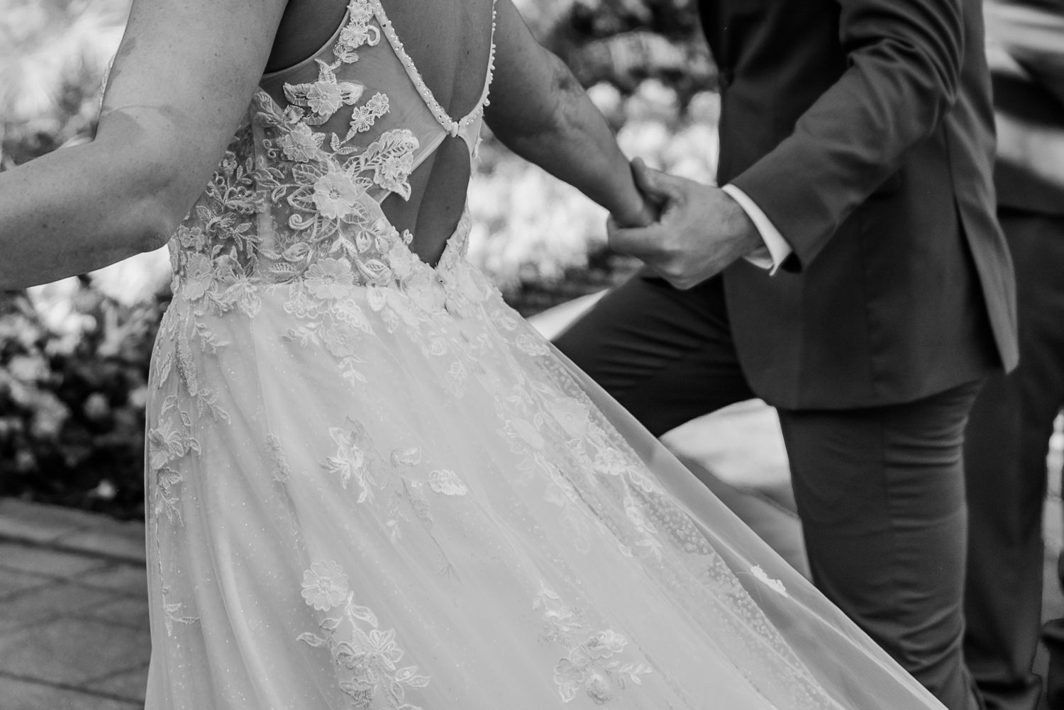 Black and white close-up of a wedding dress with delicate lace details as couple holds hands during ceremony.