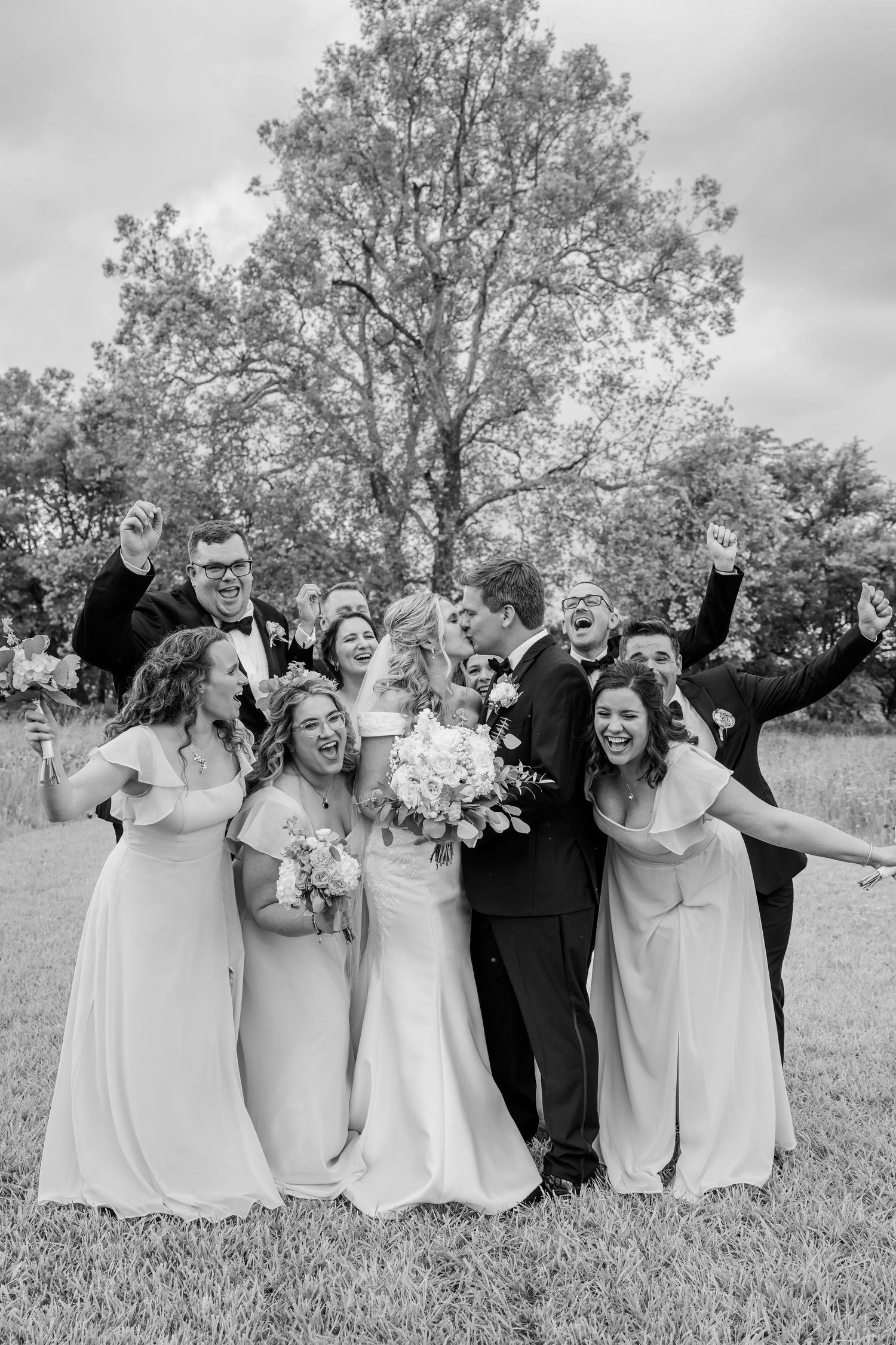 Wedding party in black and white dresses poses playfully in a grassy field with trees in the background.