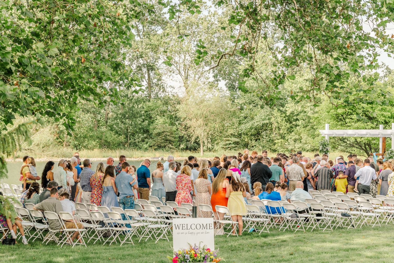 Panoramic view of outdoor wedding ceremony guests seated in white folding chairs on a lush green lawn.