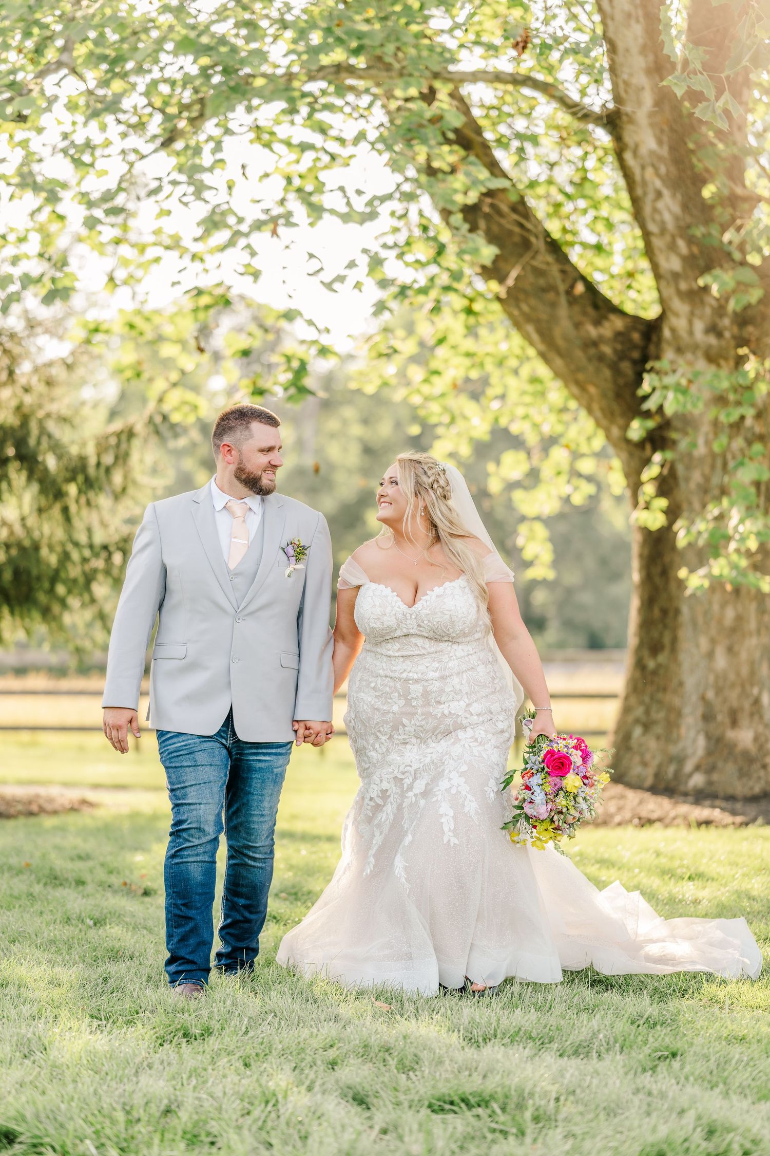 A couple in wedding attire walk hand in hand through a sunlit park with green grass and trees in the background.