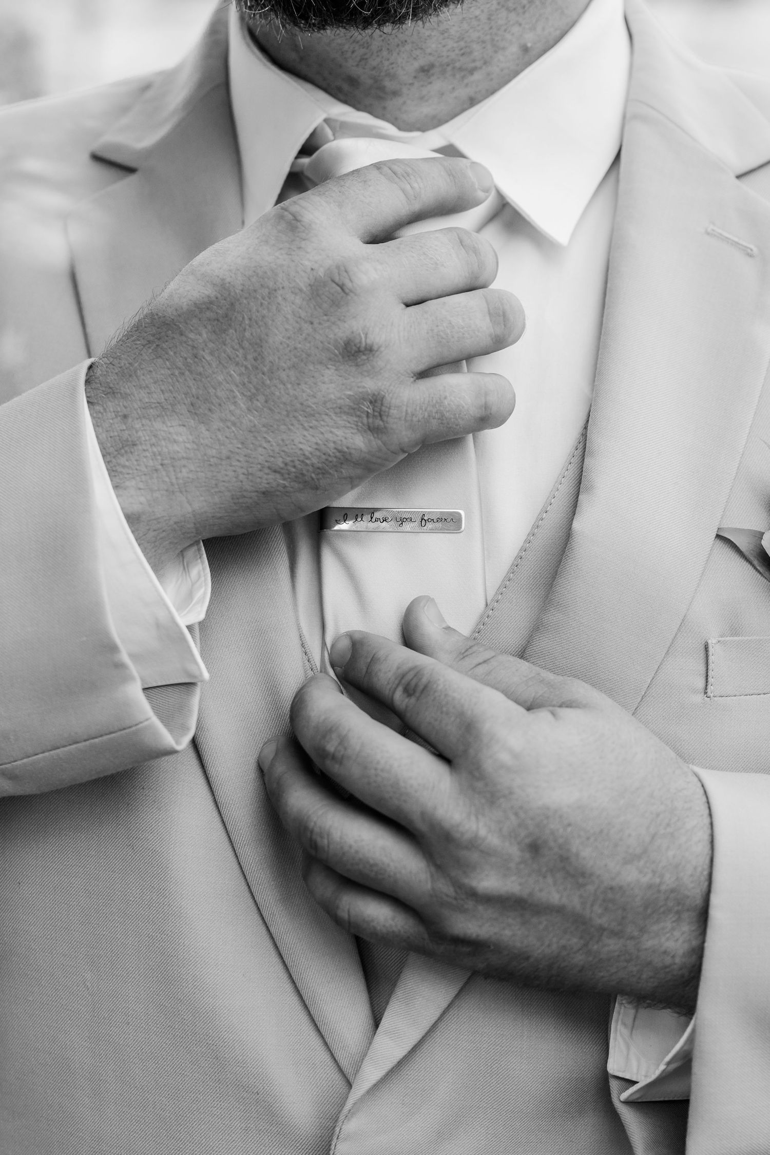 Close-up black and white photo of hands adjusting a light colored tie and suit jacket with a tie clip.
