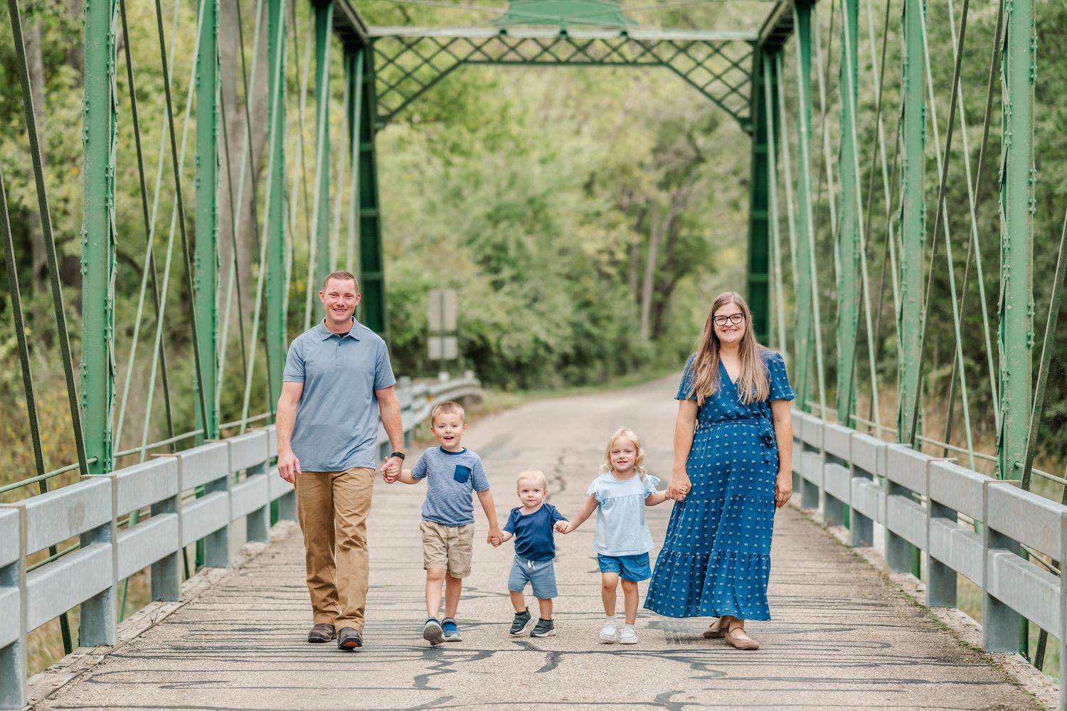 Family of five walking together and holding hands on a green metal bridge in a natural outdoor setting.