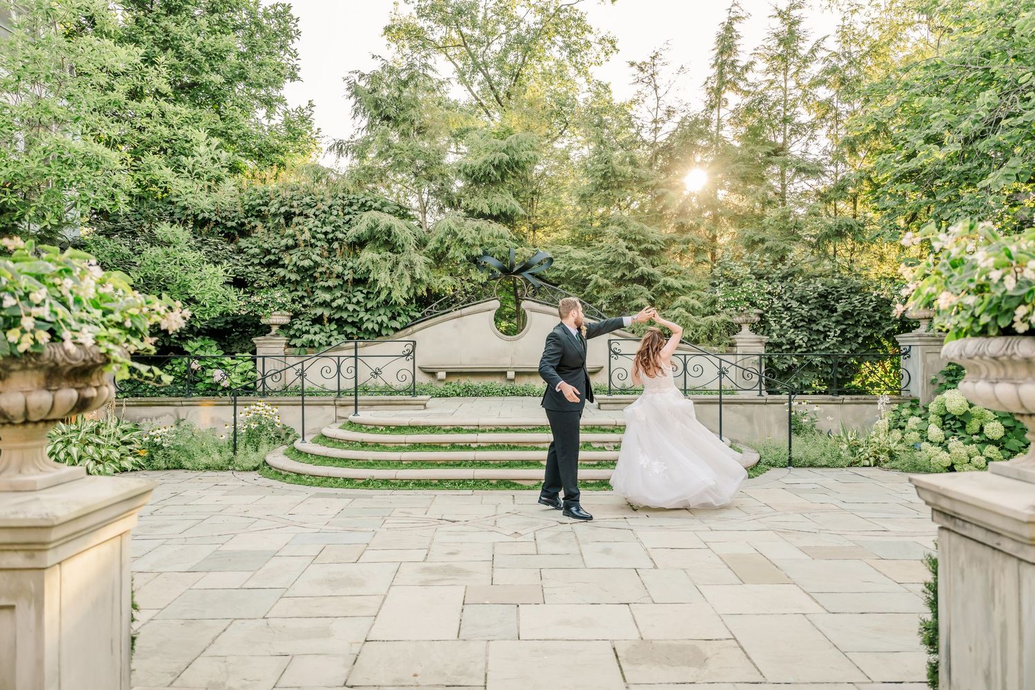 A couple in wedding attire stands on an elegant stone patio surrounded by lush gardens and trees at sunset.