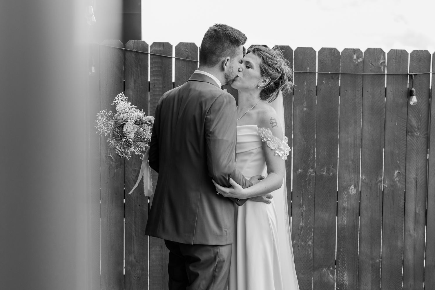 Black and white photo of a bride and groom sharing a kiss near a wooden fence, bride holding a bouquet.The Old Barns at Dry Run Farms.