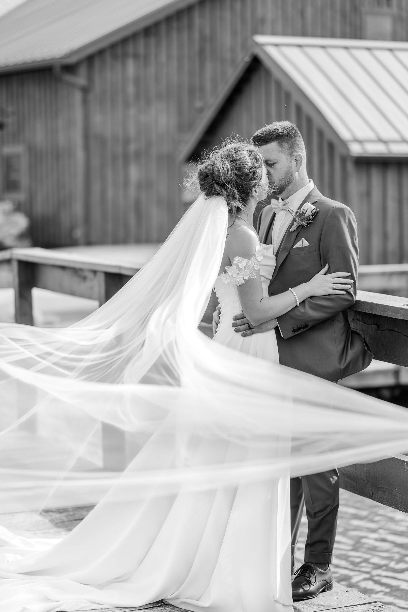 Black and white photo of bride and groom sharing a kiss with veil flowing in the wind on a wooden bridge.The Old Barns at Dry Run Farms Ohio