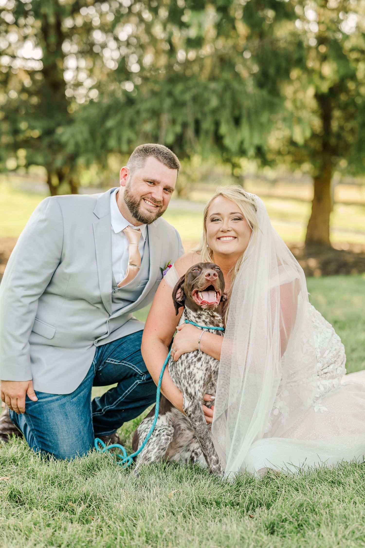 A couple in wedding attire sits on grass with their chocolate lab dog during an outdoor photo session.