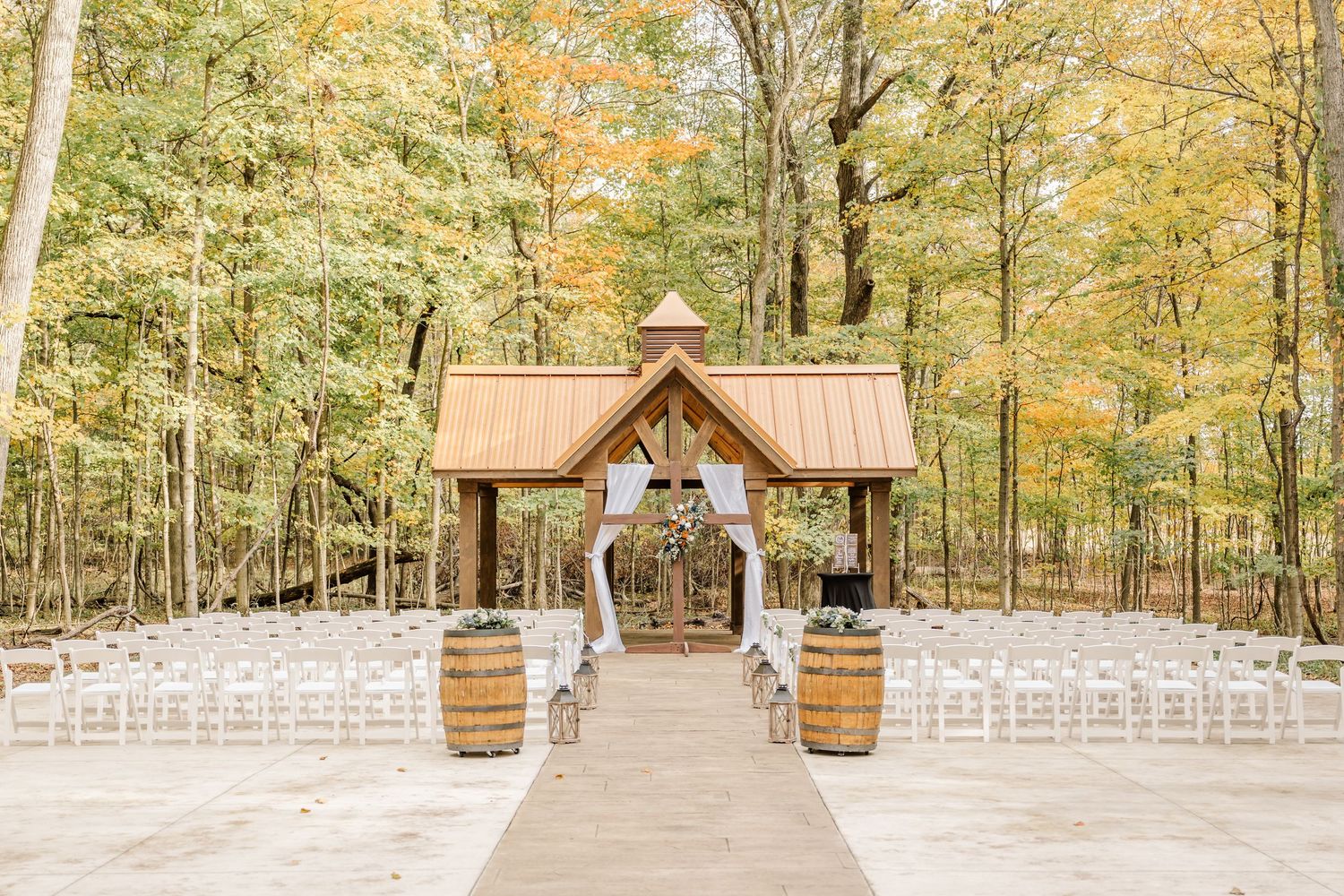A beautiful outdoor wedding ceremony setup in a wooded area with white chairs and fall foliage. Four Seasons Barn Wedding & Event Venue
