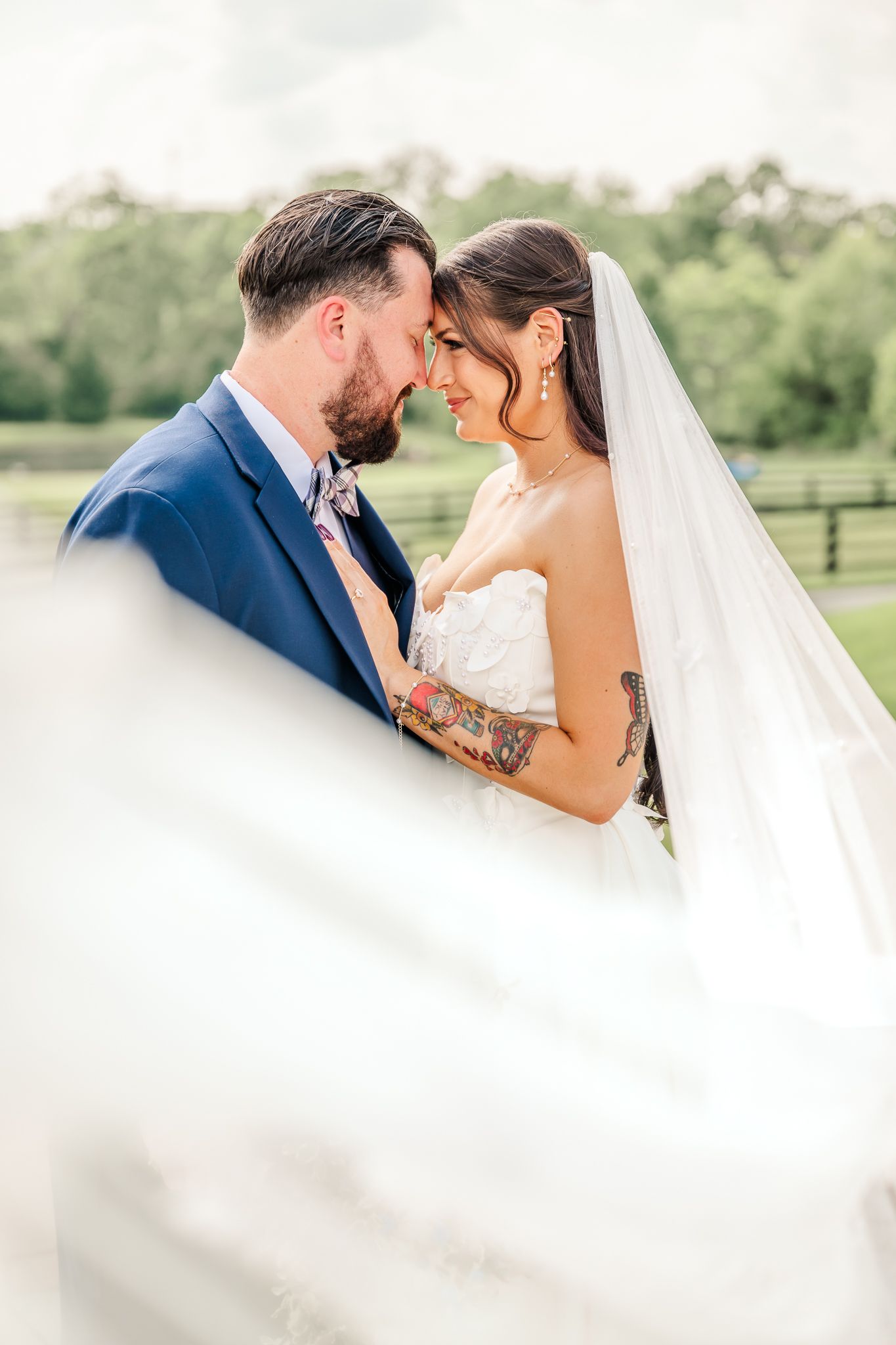 A bride & groom share an intimate touch moment, bride in lace gown with veil flowing, groom in blue suit outdoors. Canopy Creek Farm