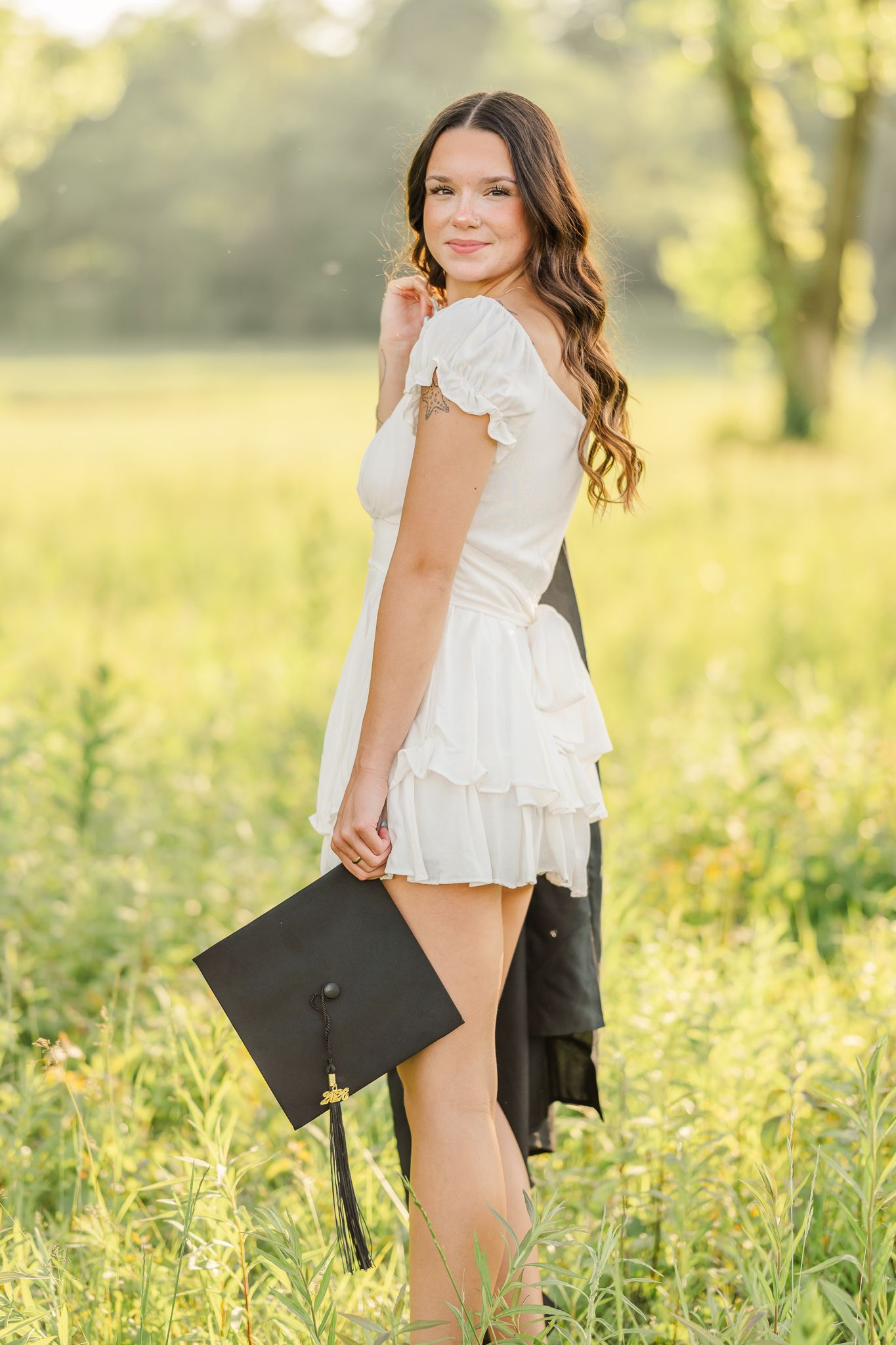 Young woman in white ruffled dress holding black graduation cap standing in sunny green field. Alum Creek Beach State Park in Lewis Center.