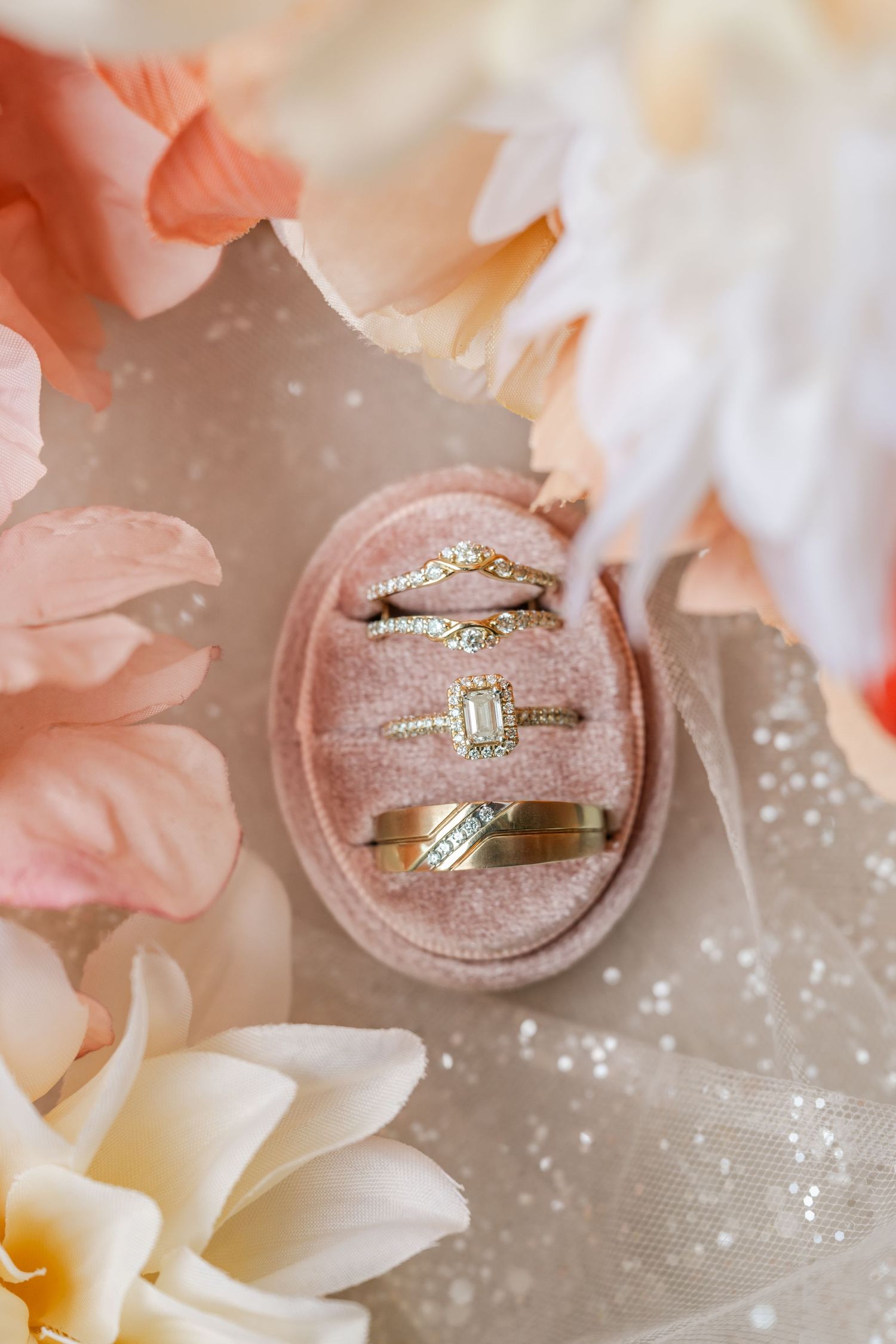 Gold wedding rings and a diamond engagement ring in a blush pink velvet ring box surrounded by peach and white flowers.