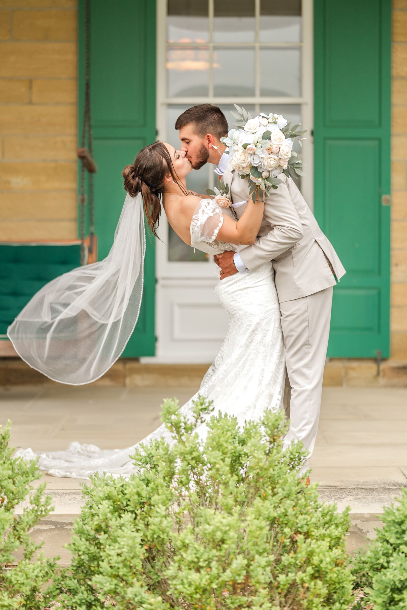 Bride and groom share a romantic dip kiss outdoors, bride in lace gown with flowing veil, groom in gray suit. Brynn Du Mansion Granville, OH