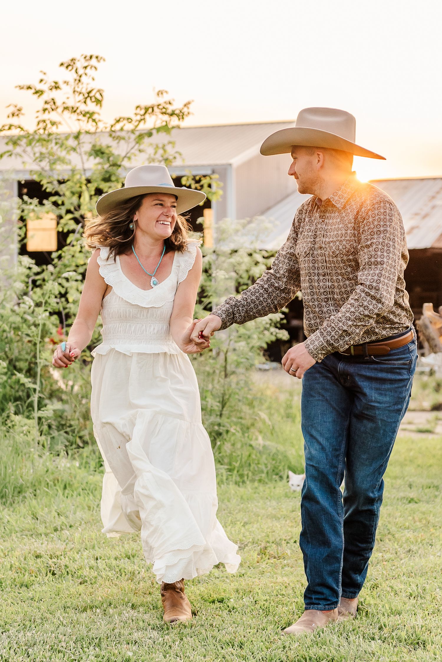 Couple in western attire dancing playfully in a grassy field at sunset.