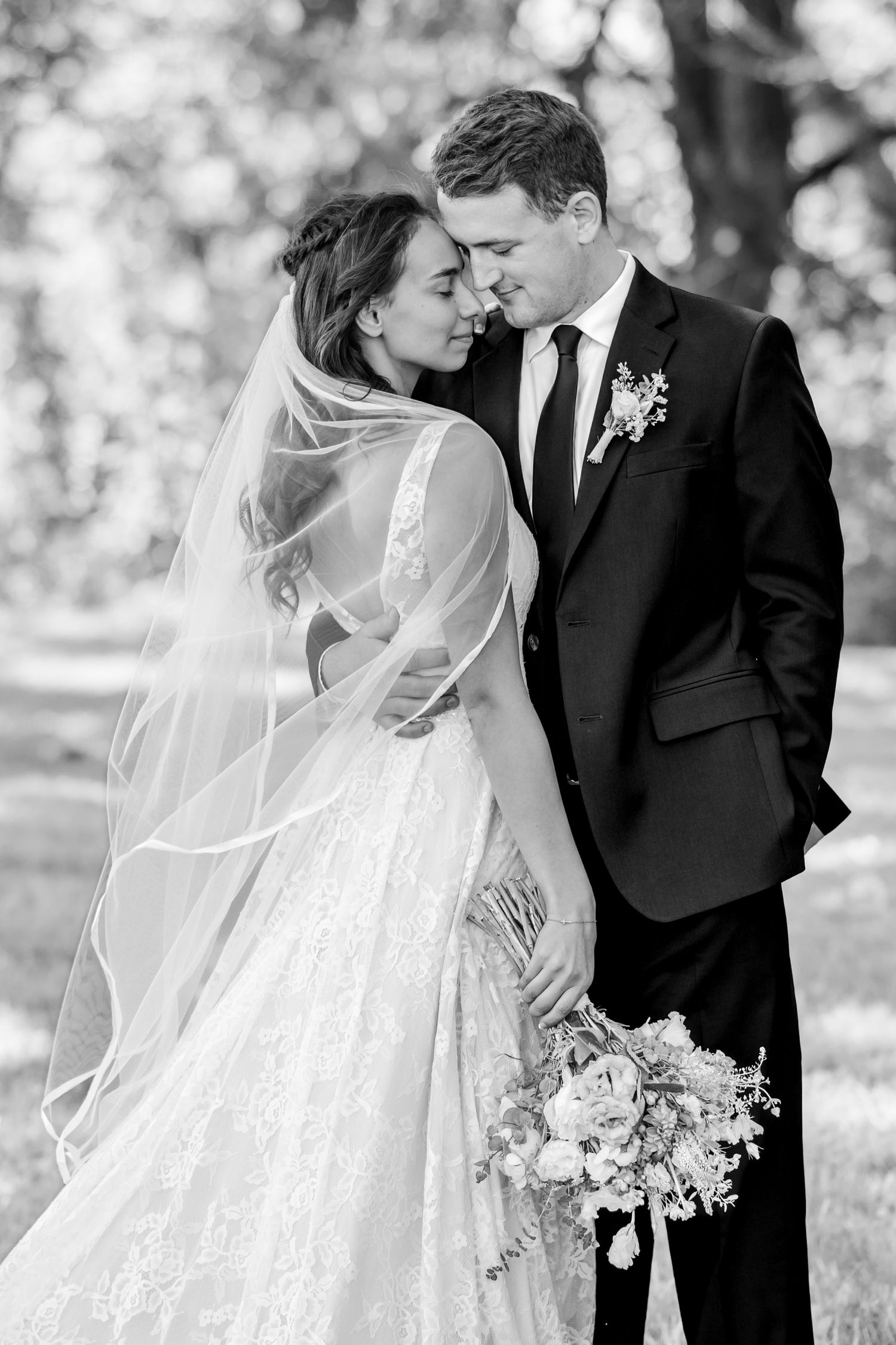 Black and white photo of a bride in a lace gown and veil embracing her groom in a suit with boutonniere outdoors, holding a bouquet.