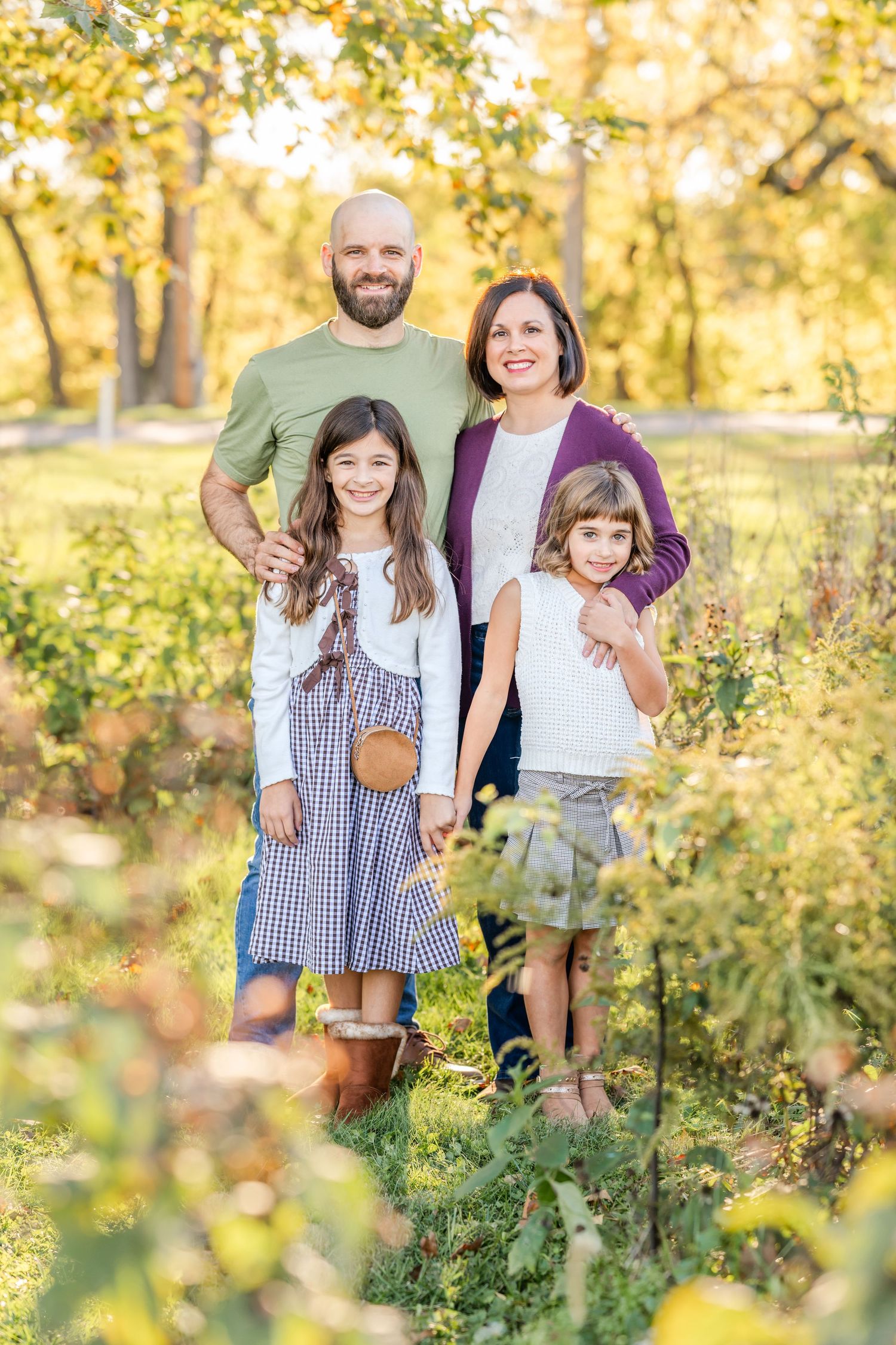 Smiling family of four posing outdoors in a sunlit autumn field, with two girls standing in front of their parents among golden foliage.