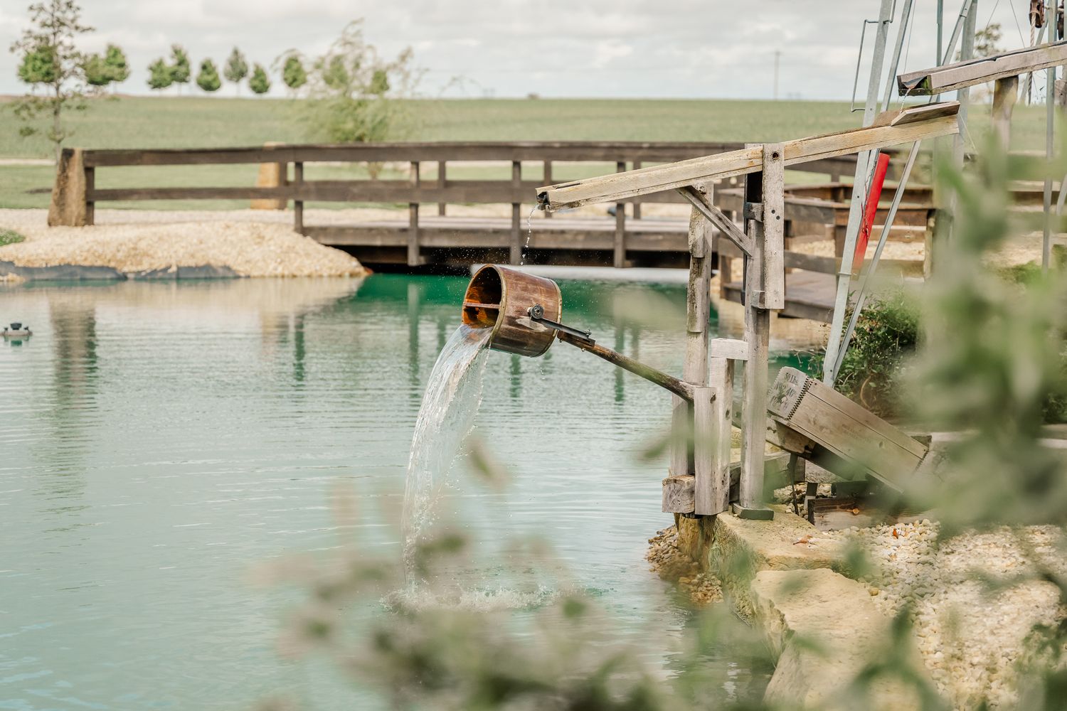 A wooden pipe pours water into a serene green pond beside a rustic bridge surrounded by lush greenery.The Old Barns at Dry Run Farms.