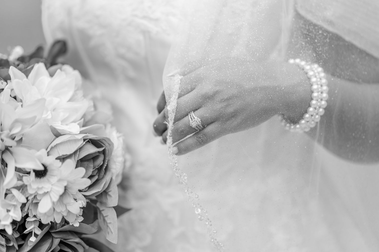 Black and white photo of a bride in a lace gown and veil holding a floral bouquet, wearing a pearl bracelet and a ring.