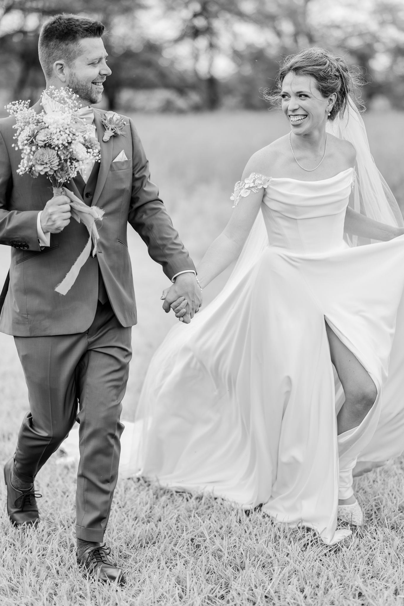 Black and white photo of smiling bride and groom holding hands walking through a field on their wedding day.