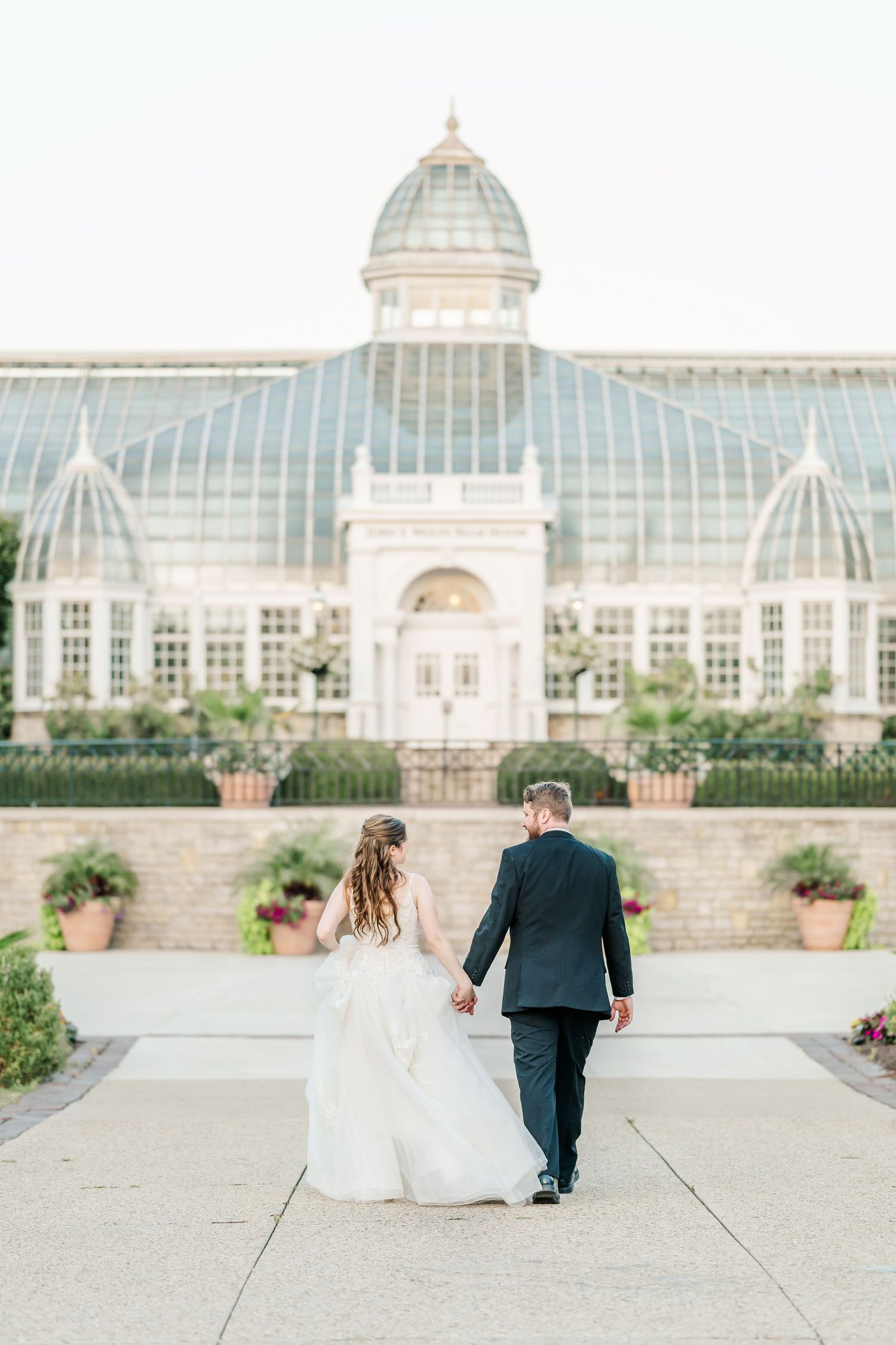 Wedding couple walking away from a grand conservatory building with ornate glass architecture.
