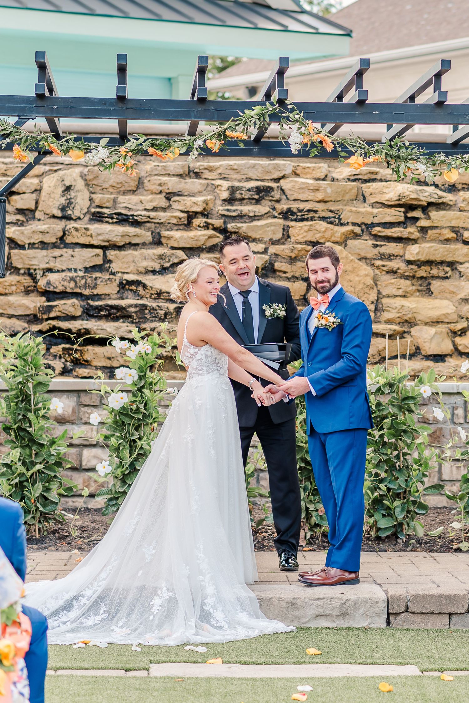 A wedding ceremony takes place in front of a rustic stone wall with greenery and flowers decorating the outdoor venue.