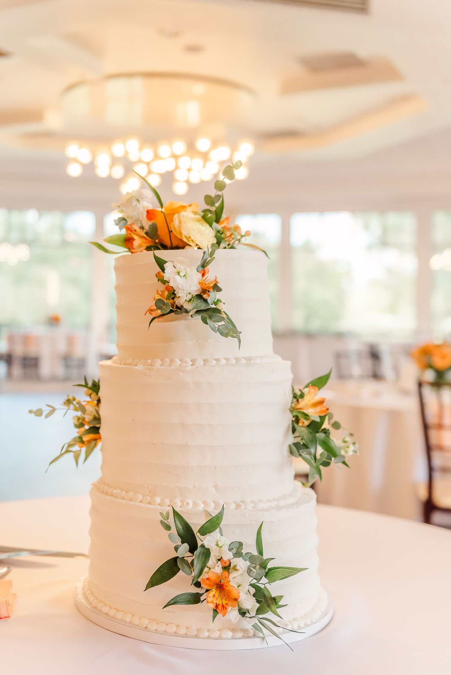 White tiered wedding cake decorated with orange and green floral accents sits elegantly on display at reception venue.