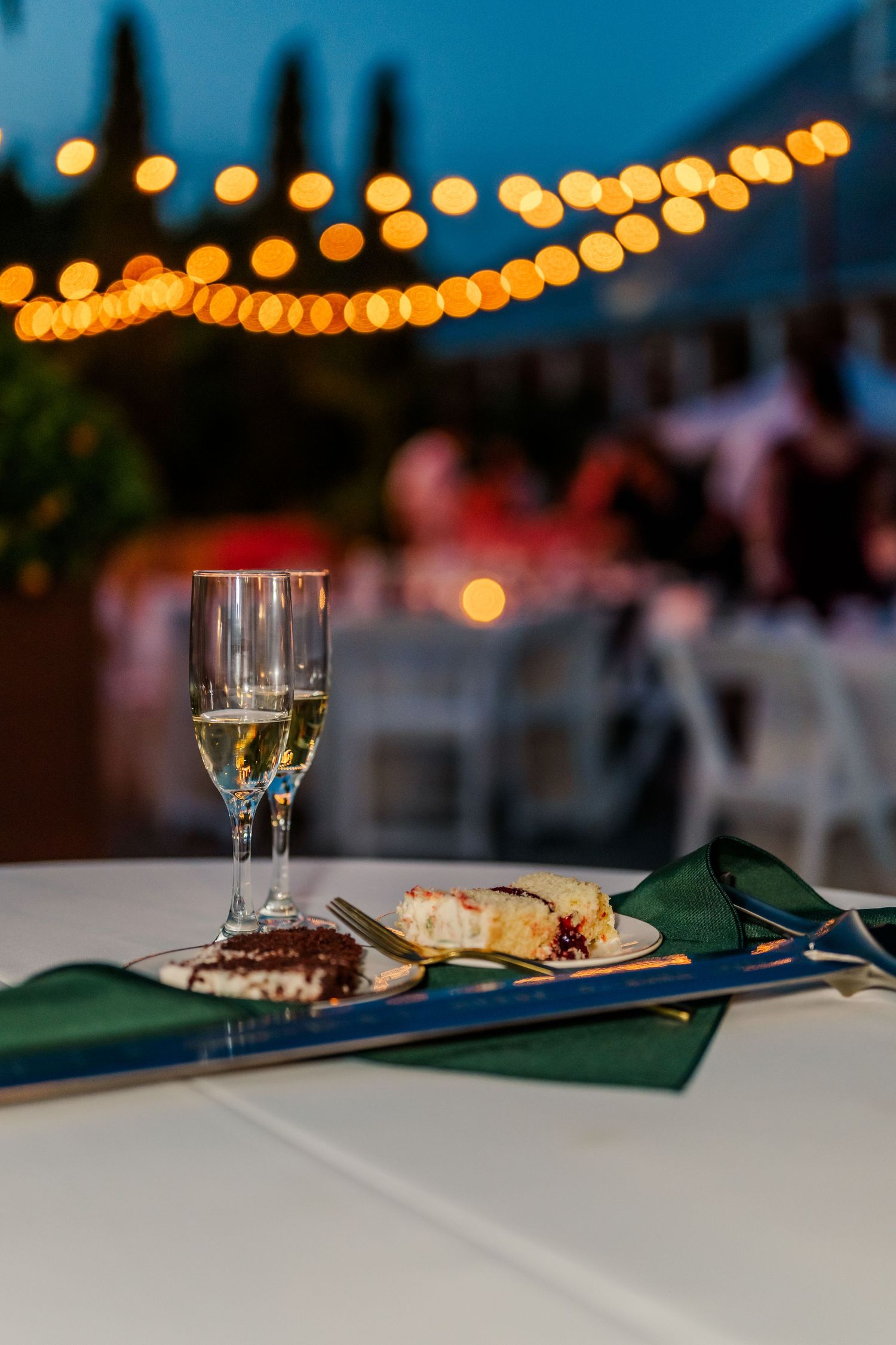 Wedding reception dinner plates and wine glasses on white tablecloths under glowing string lights at dusk.