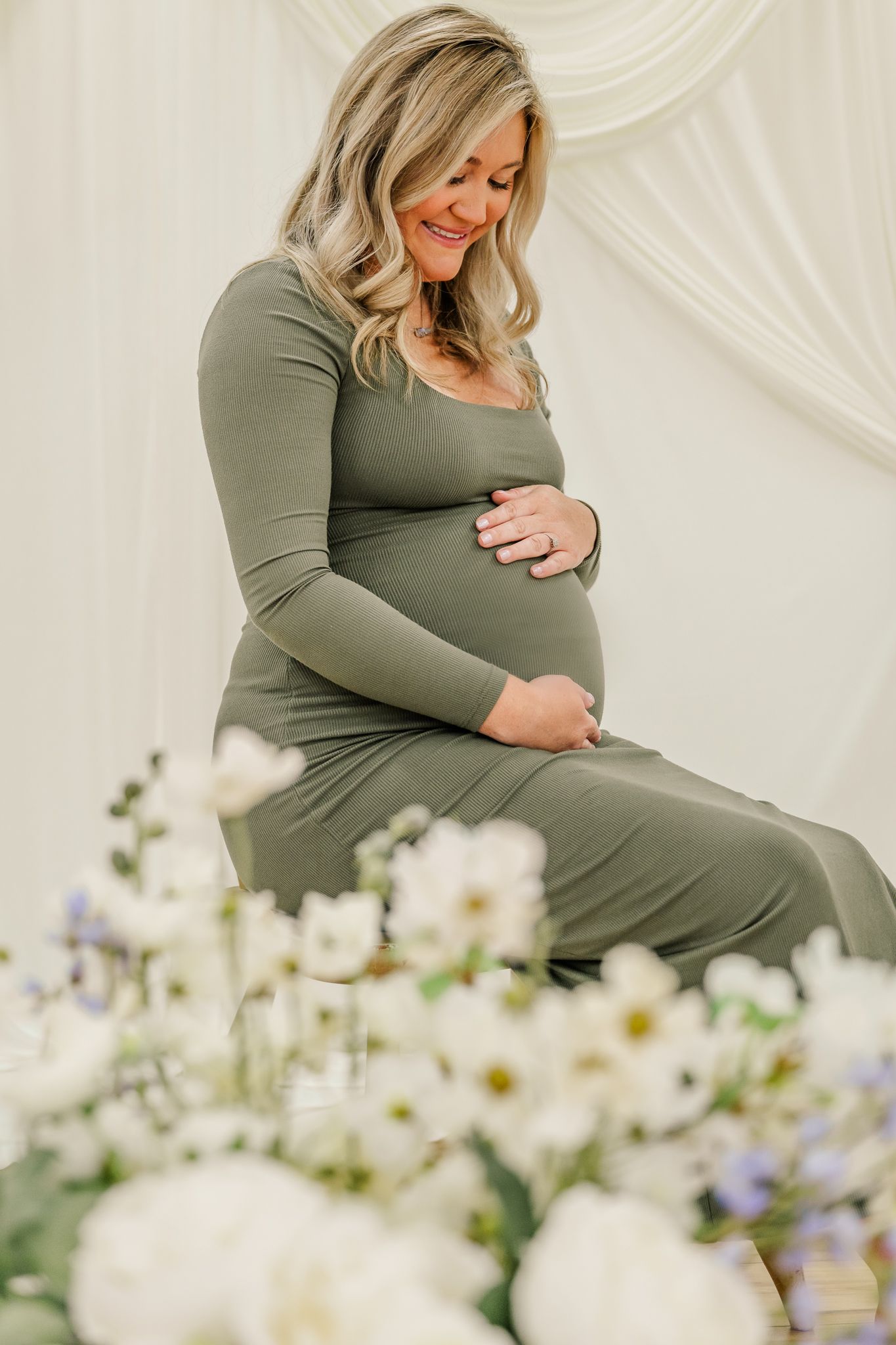 Pregnant woman in olive green maxi dress cradling baby bump, surrounded by white flowers at a maternity photo shoot.